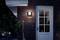 Die Außenansicht eines Hauses bei Nacht, beleuchtet von einer Wandleuchte über einer weißen Tür mit Glaselementen, Pflanzen daneben.