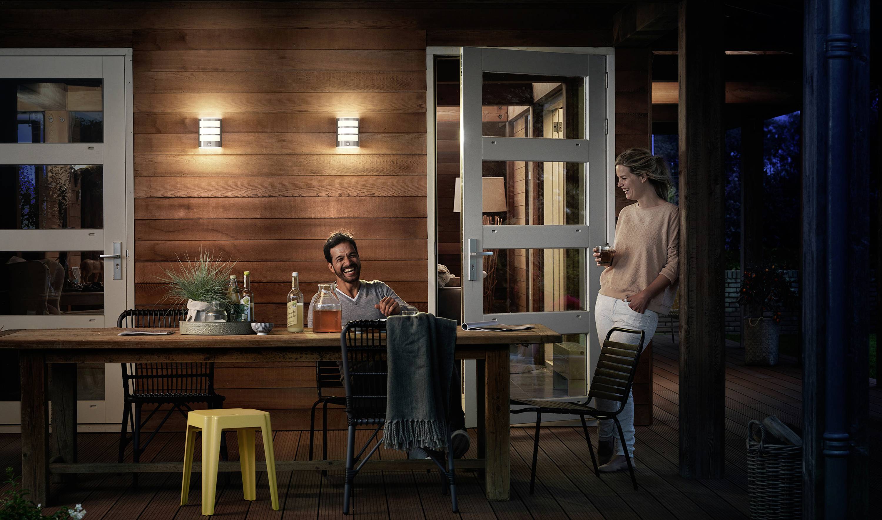 Ein Mann und eine Frau unterhalten sich entspannt auf einer Terrasse am Abend. Die Umgebung ist warm beleuchtet und ein Tisch ist gedeckt.