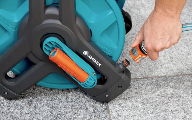 Eine Person schließt einen Gartenschlauch an eine mobile blaue Schlauchtrommel mit einem orangefarbenen Düsengriff auf einer Steinpatio-Oberfläche an.