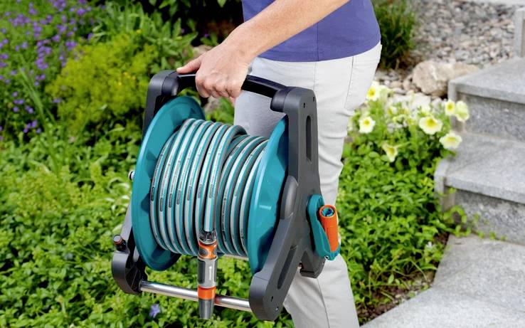 Eine Person trägt eine mobile Gartenschlauchtrommel in einem Garten mit blühenden Blumen und Grünpflanzen, neben steinernen Stufen.