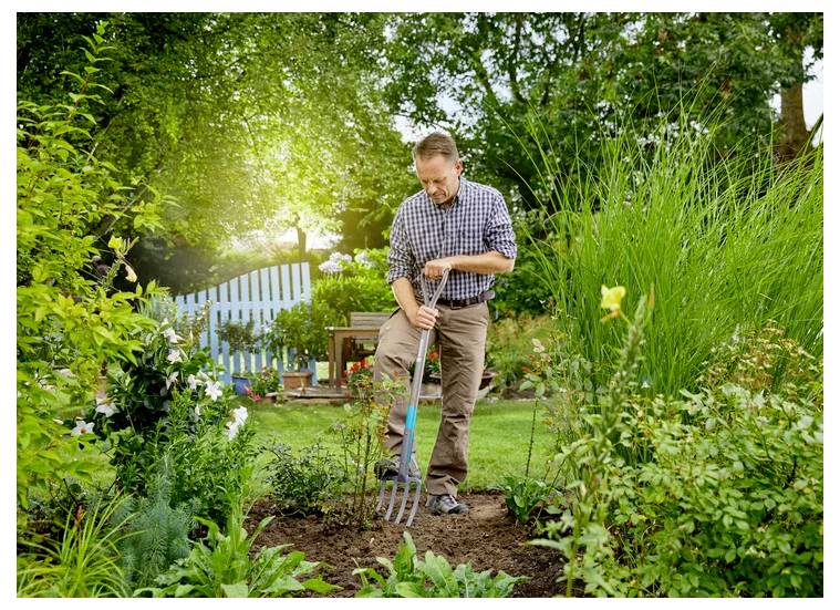 Ein Mann in einem Garten verwendet eine Mistgabel, um die Erde umzugraben, umgeben von grünen Pflanzen und einem blauen Tor im Hintergrund.