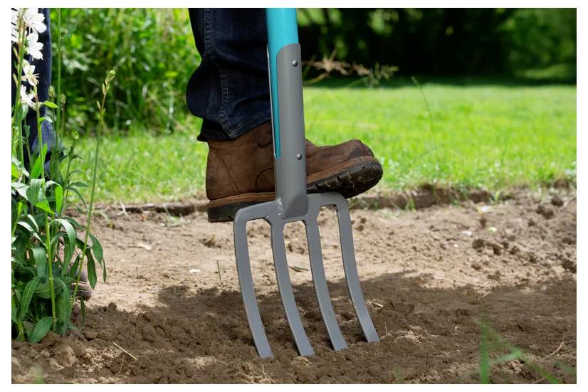 Eine Person in Stiefeln verwendet eine Grabgabel, um Erde in einem Garten umzugraben, umgeben von Gras und Pflanzen, was auf eine Gartenarbeit hindeutet.