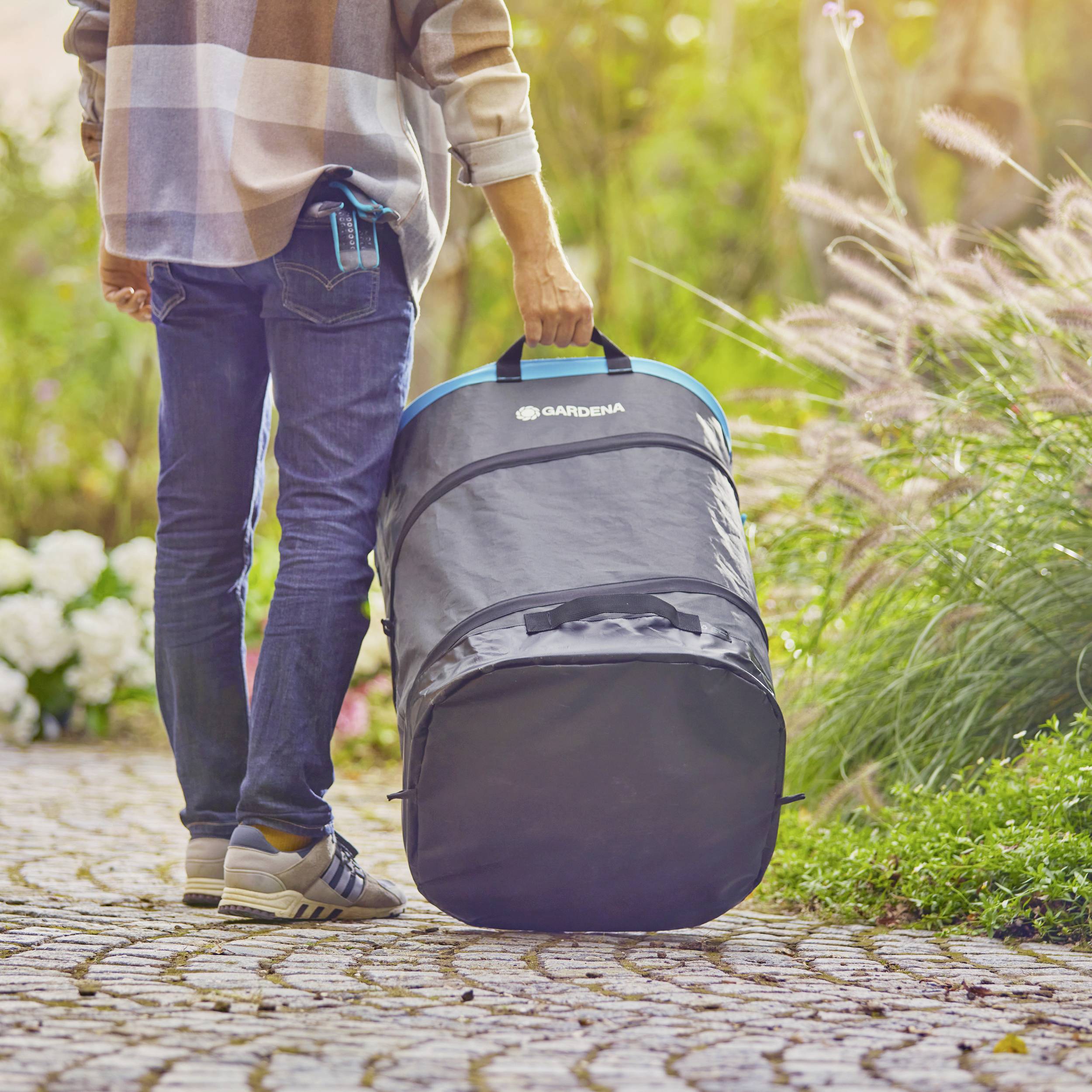 Eine Person mit kariertem Hemd und Jeans hält eine große Tasche von Gardena auf einem gepflasterten Weg in einem Gartenbereich.
