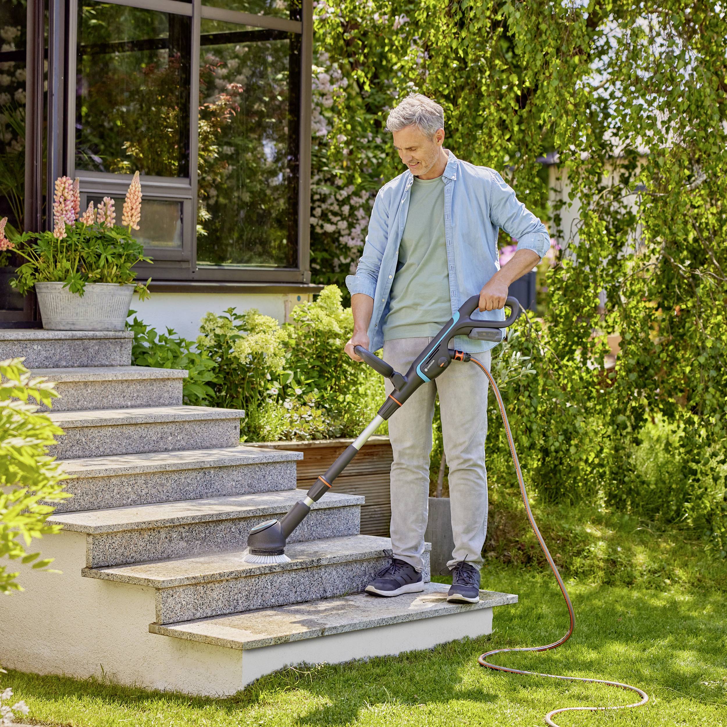 Ein Mann verwendet einen Staubsauger, um eine Außentreppe im Garten sauber zu machen. Im Hintergrund sind Pflanzen zu sehen.