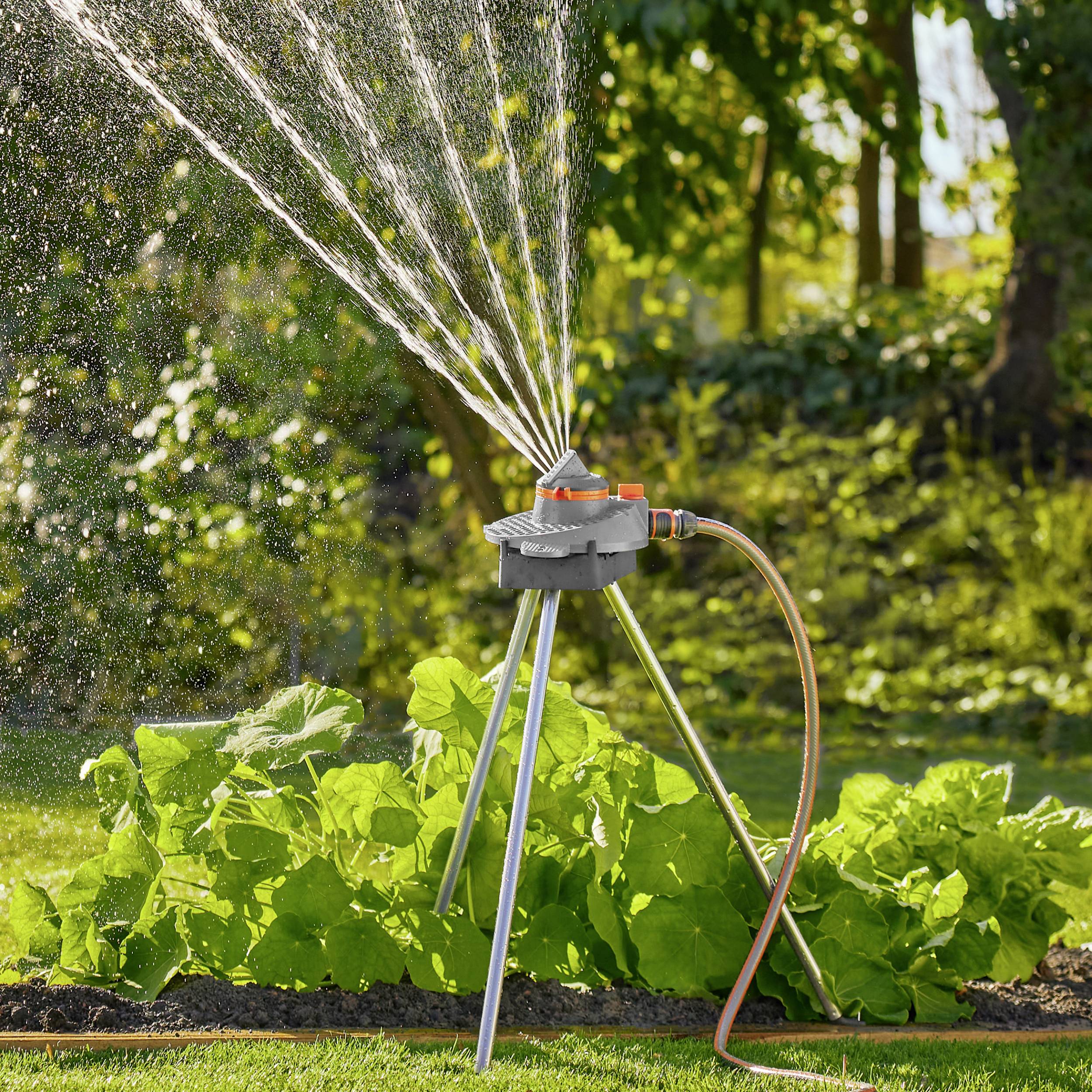 Ein Gartensprinkler verteilt Wasserstrahlen über einen gepflegten Rasen, umgeben von Büschen, unter sonnigem Himmel.