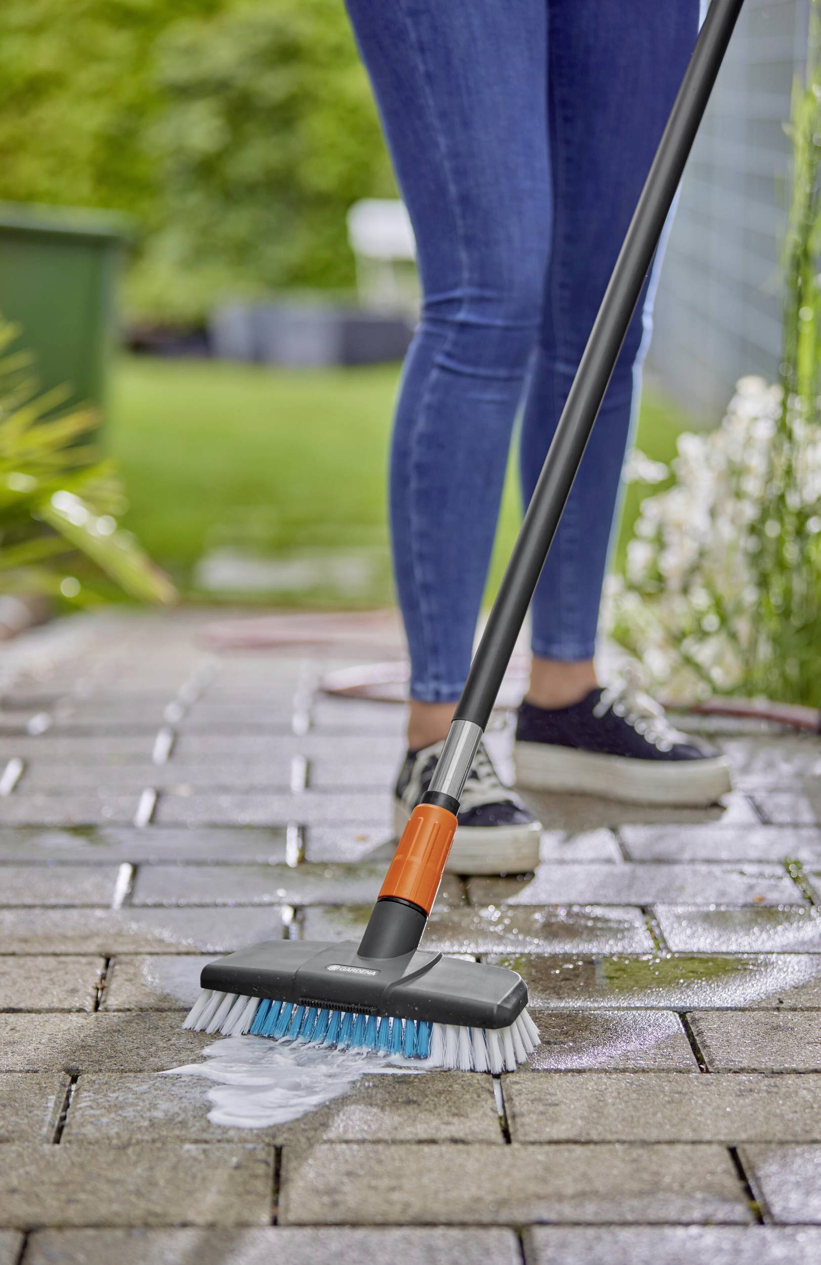 Person in Jeans reinigt mit Bürste und Wasser den Gehweg. Im Hintergrund sind Gartenpflanzen sichtbar.