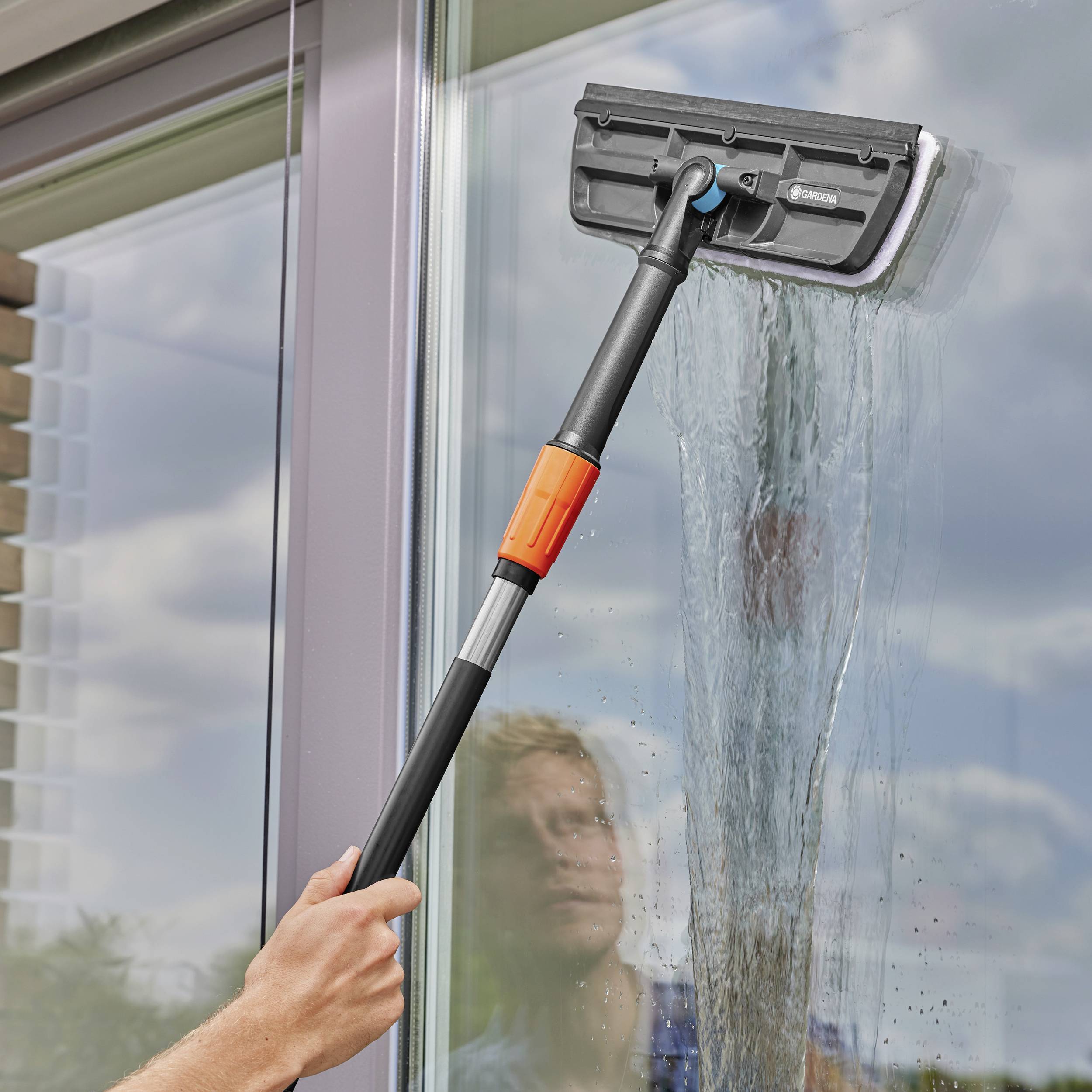 Eine Person reinigt ein großes Fenster mit einem Teleskop-Fensterwischer. Im Hintergrund ist der blaue Himmel mit Wolken zu sehen.