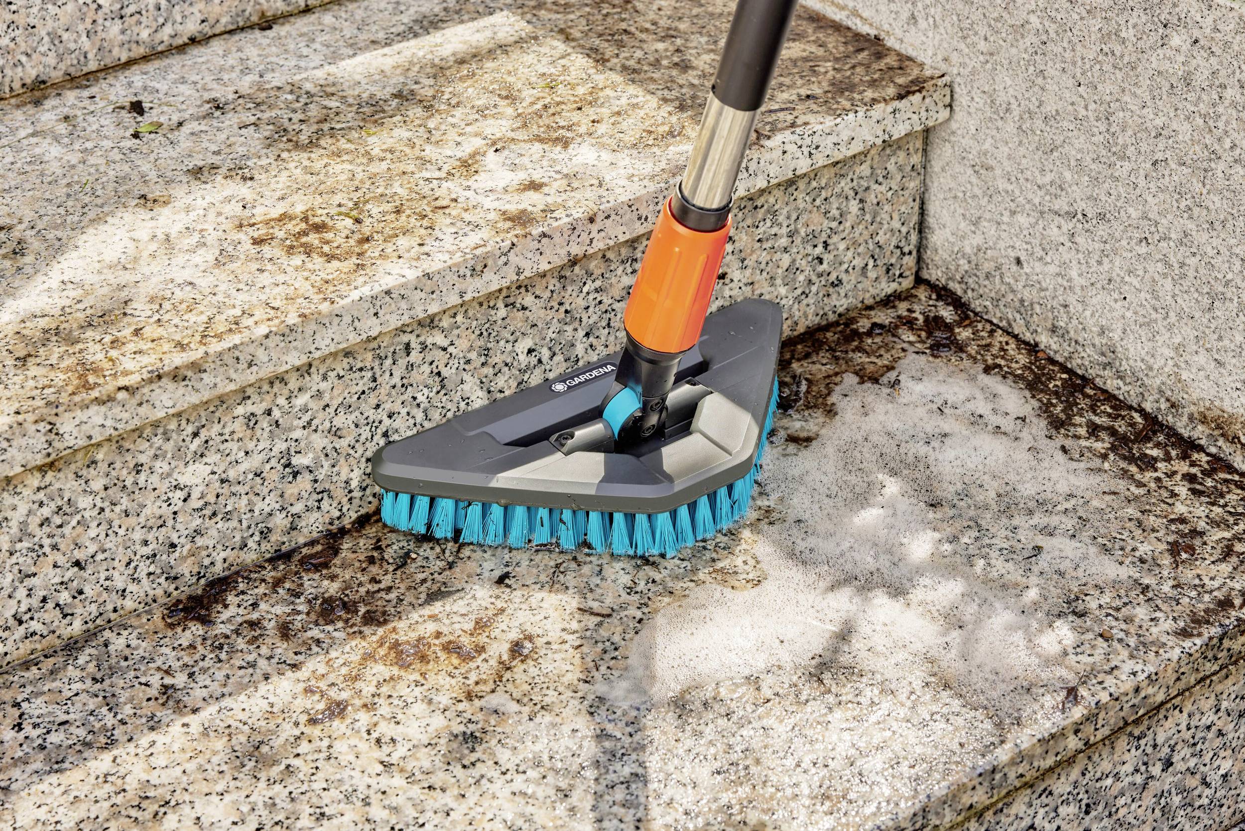 Eine Treppe aus Stein wird mit einer Bürste gereinigt. Die Bürste hat blaue Borsten und wird von einem langen Griff gehalten. Schmutz und Seifenlauge sind sichtbar.