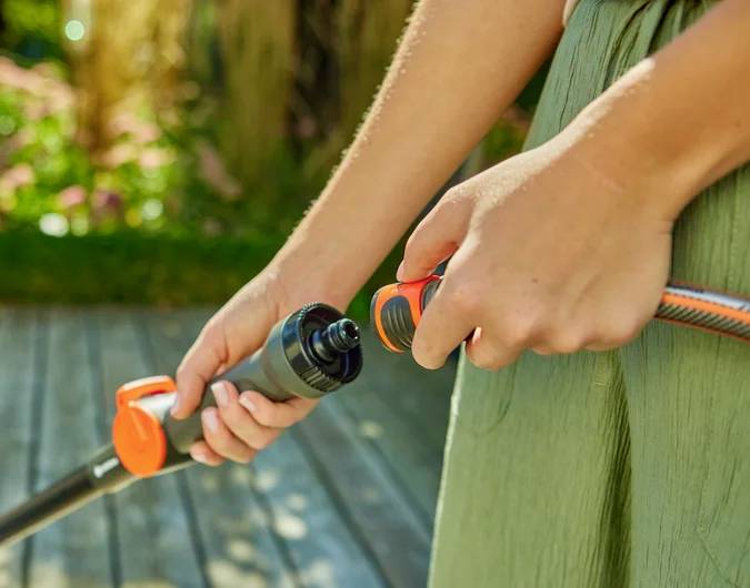 Eine Person, die einen Gartenschlauch mit einem orangefarbenen Aufsatz auf einer sonnigen Holzterrasse anschließt, was die Nutzung von Wasser im Freien zeigt.
