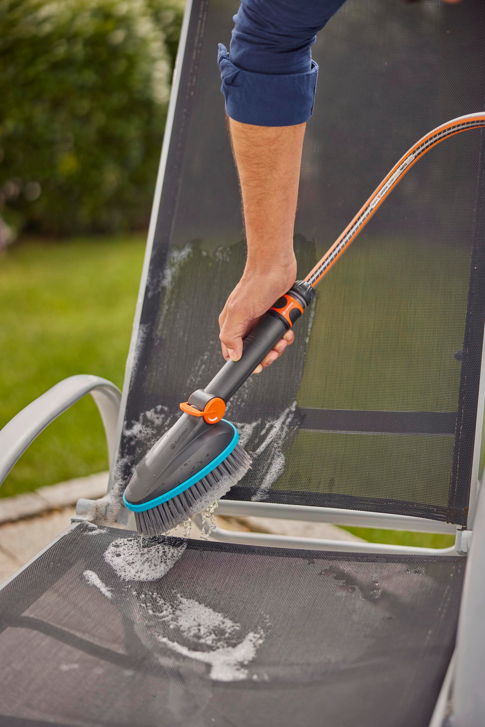 Eine Person reinigt mit einer Bürste einen Gartenstuhl im Freien. Schaum bedeckt die Sitzfläche des Stuhls.