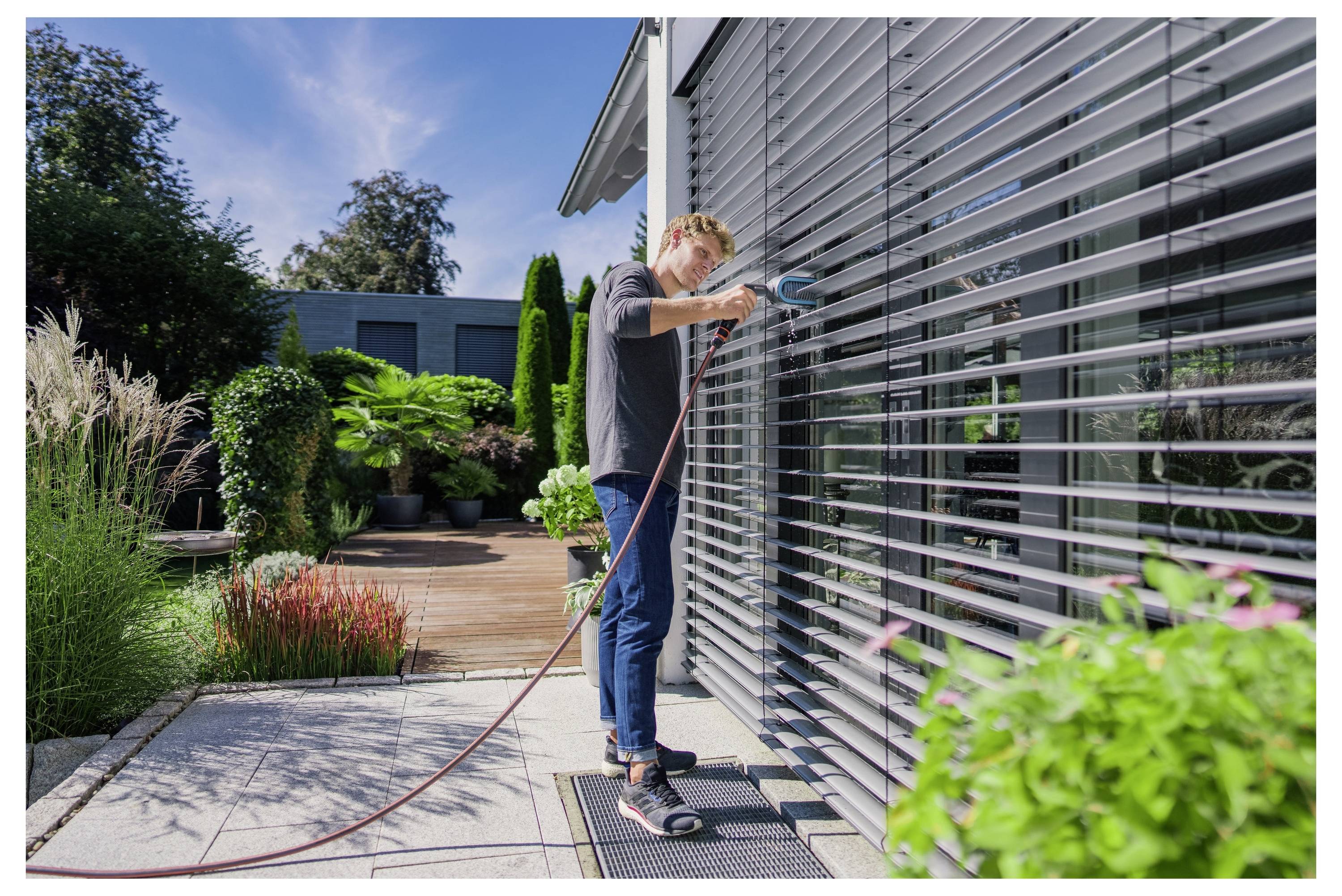 Eine Person, die große Fensterläden mit einem Schlauch außerhalb eines modernen Hauses reinigt, umgeben von einem Garten- und Terrassenbereich.