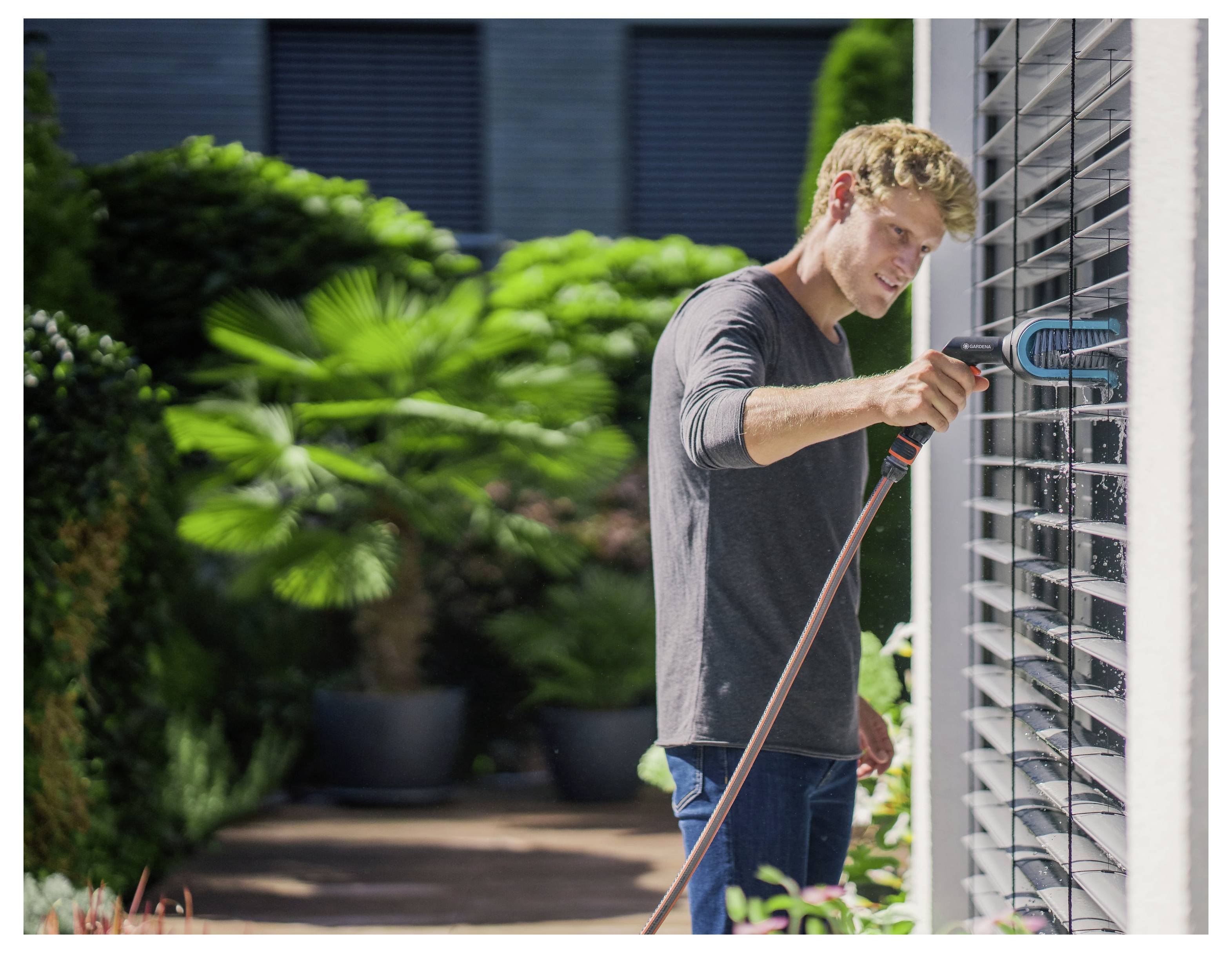 Eine Person reinigt Außenjalousien mit einer Bürste und einem Schlauch. Im Hintergrund sind üppig grüne Pflanzen sichtbar.