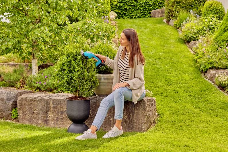 Eine Frau sitzt auf einer Steinmauer in einem üppigen Garten und bewässert Pflanzen mit einem blauen Schlauch. Sie wirkt zufrieden, umgeben von Grün und Blumen.