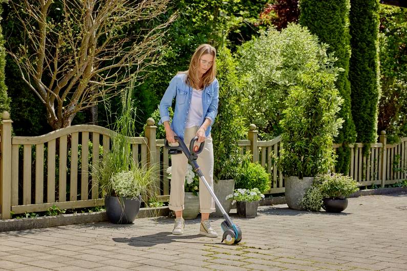 Eine Frau benutzt einen elektrischen Rasentrimmer, um Unkraut zwischen Pflastersteinen in einem Garten zu schneiden. Im Hintergrund sind Pflanzen und ein Holzzaun zu sehen.