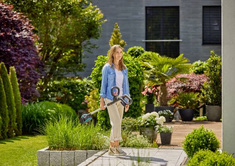 Eine Frau steht in einem gut gepflegten Garten und hält einen tragbaren Rasentrimmer. Der Garten ist voller grüner Pflanzen und bunter Blumen.