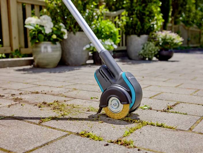 Ein Elektro-Fugenreiniger mit rundem Drahtbürstenkopf entfernt Moos und Unkraut von Pflastersteinen in einem Gartenbereich.
