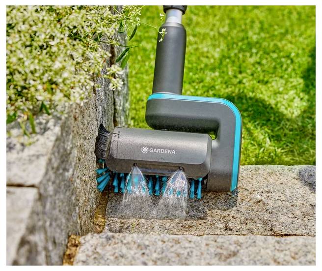 Eine tragbare Bürste von Gardena, die Wasser auf eine Steinmauer spritzt, um sie zu reinigen. Sie ist von Grünpflanzen und Gras umgeben.