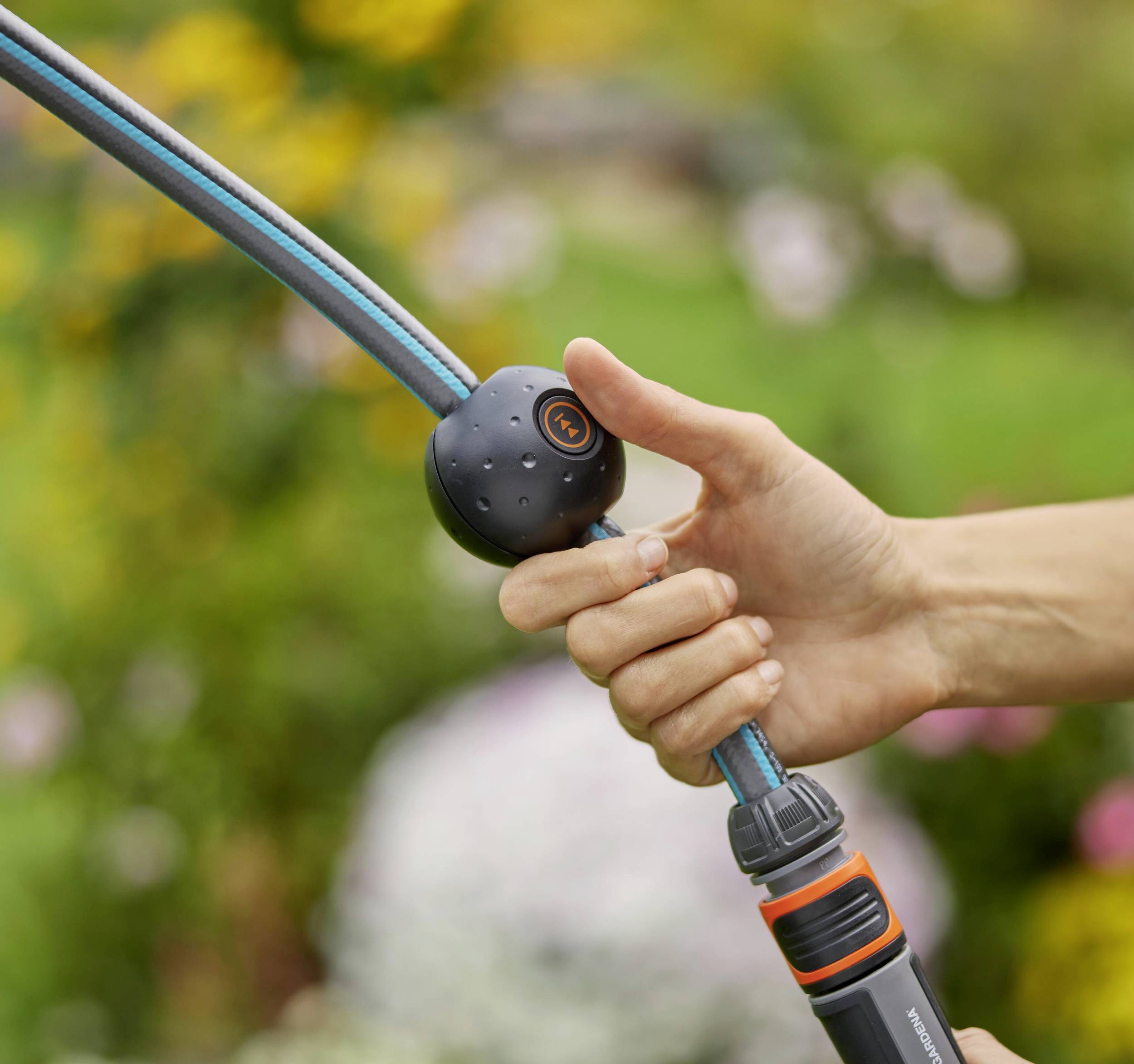 Eine Hand hält einen Gartenschlauch mit Düse. Der Garten im Hintergrund zeigt unscharfe Blumen, was eine frühlingshafte Umgebung andeutet.