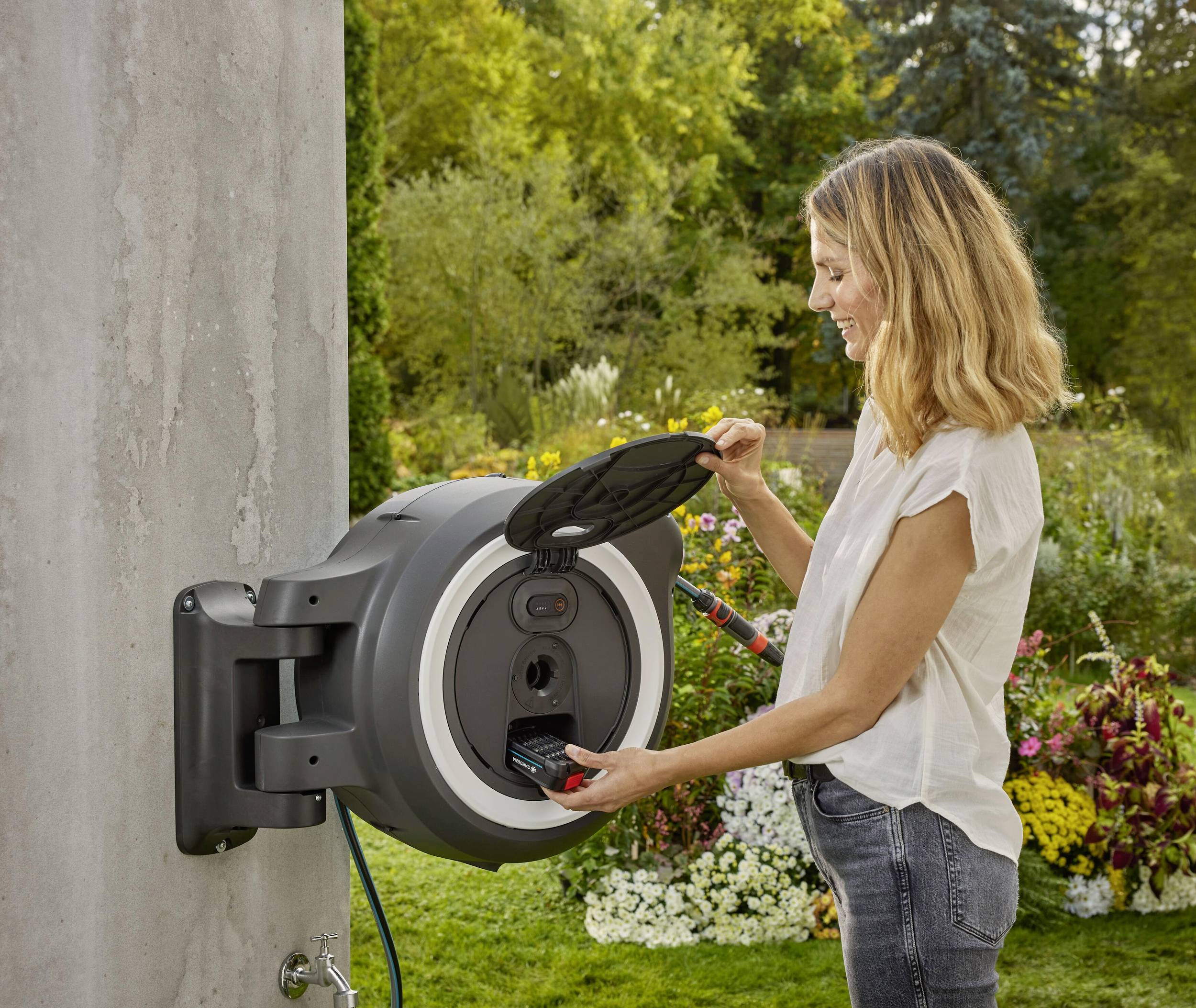 Eine Frau bedient ein modernes Gartenschlauchsystem an einer Wand in einem blühenden Garten. Sie hebt den Deckel des Systems an.