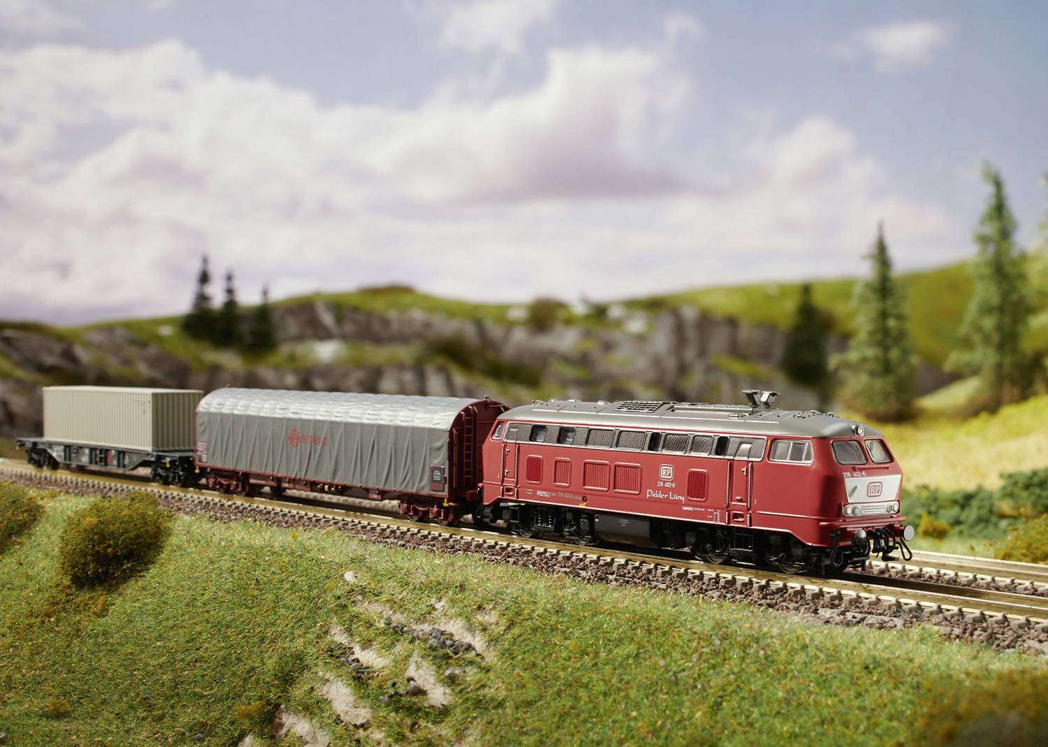 Eine rote Modelllokomotive fährt auf einer Landschaft mit Wiese und Bäumen im Hintergrund auf einer Modelleisenbahn.