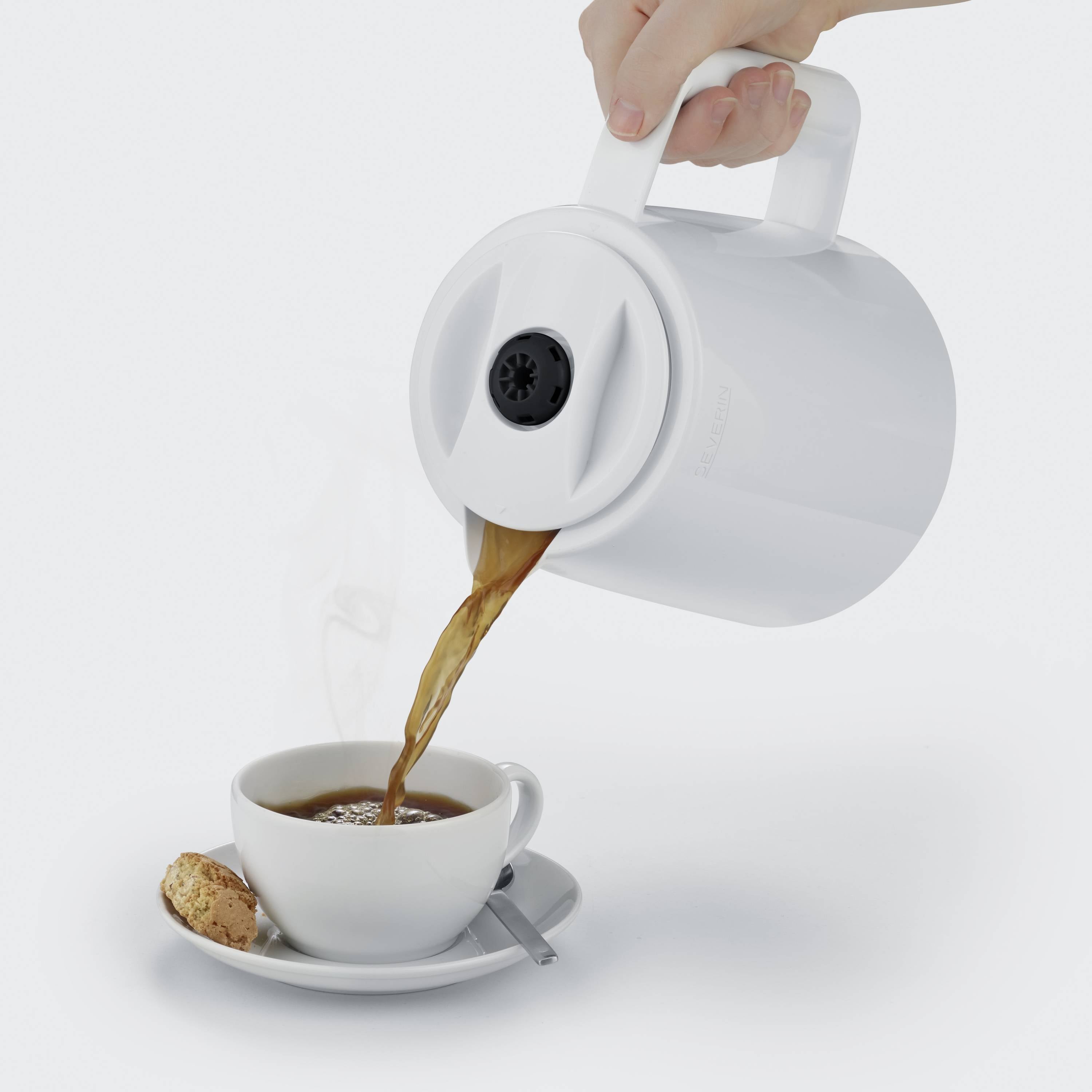 Eine Hand gießt Kaffee aus einer weißen Thermoskanne in eine Tasse mit einem Keks und einem Löffel auf der Untertasse.
