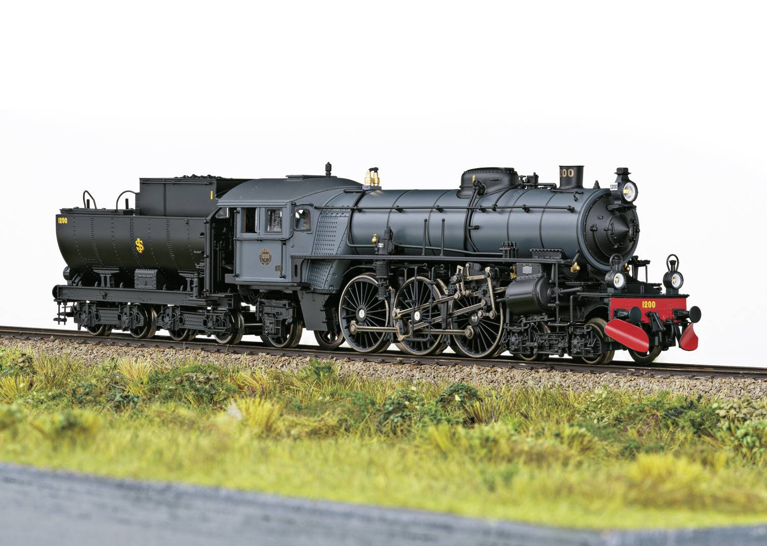 Dampflok-Modell auf einem Gleis, detailliert dargestellt vor einem weißen Hintergrund, umgeben von Gras. Lokomotive in grau mit roten Details.