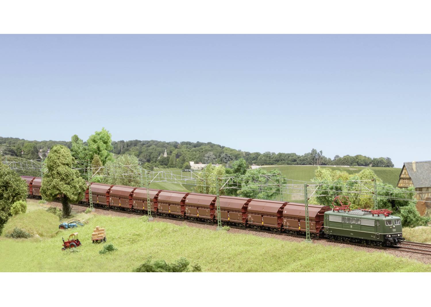 Ein grüner Güterzug fährt durch eine ländliche Landschaft mit Bäumen und Wiesen. Im Hintergrund sind Hügel und ein Bauernhof zu sehen.