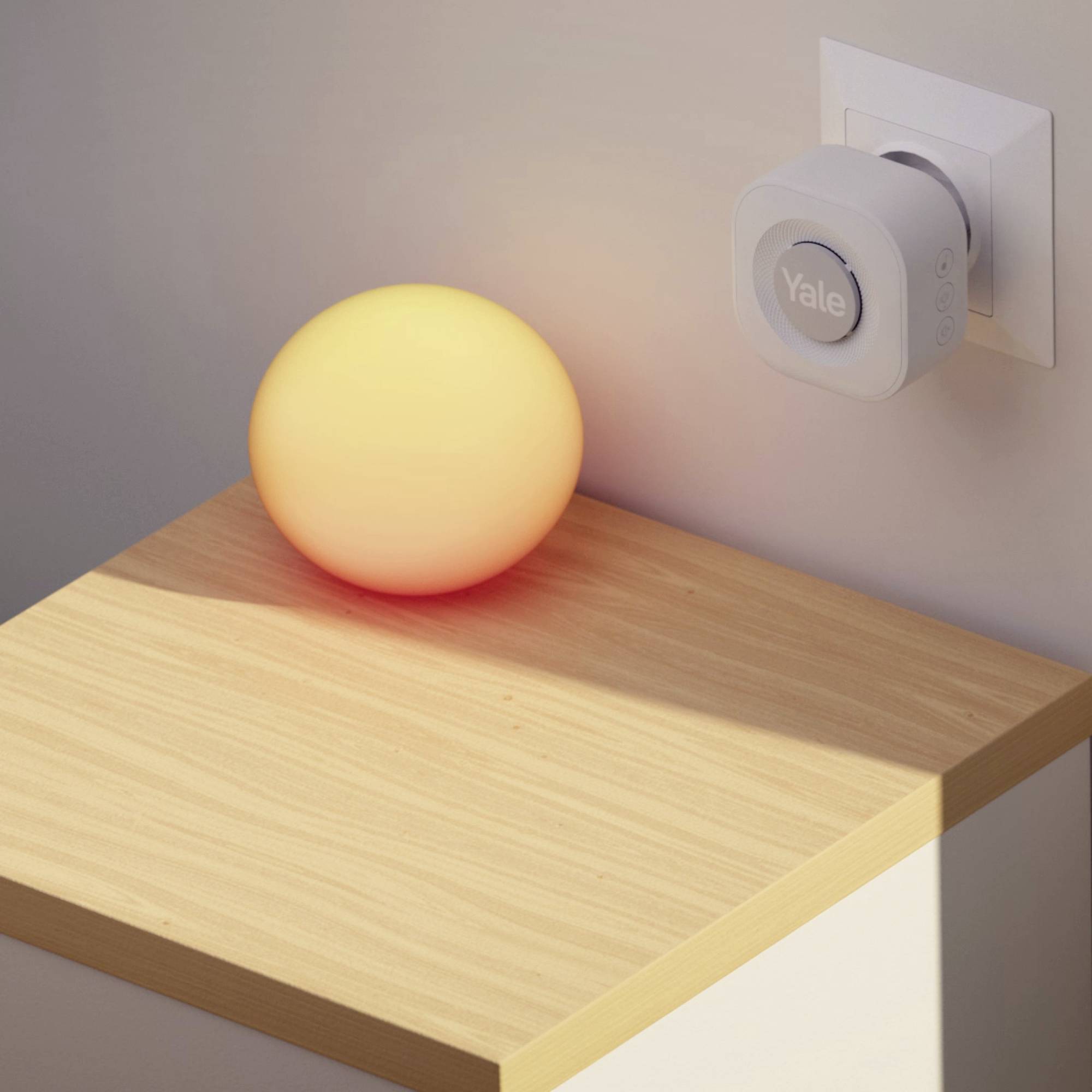 Ein runder, leuchtender, gelber Gegenstand liegt auf einem Holztisch. Eine weiße Steckdose mit grauem Gerät ist an der Wand befestigt.