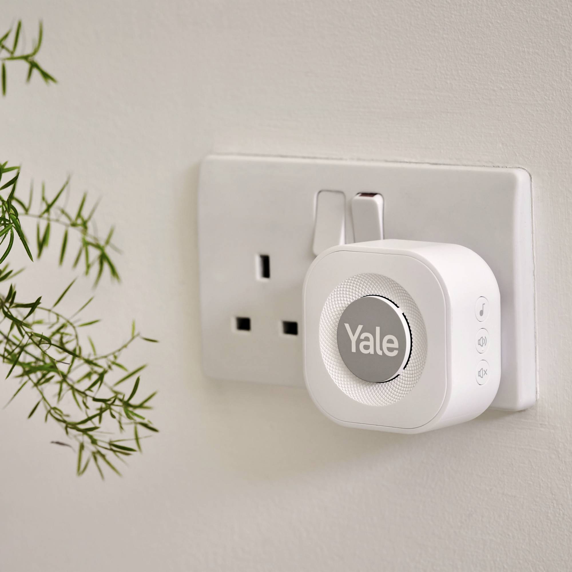 Ein weißer Smart-Stecker von Yale ist in eine Wandsteckdose eingesteckt. Im Vordergrund ist ein Zweig mit grünen Blättern zu sehen.
