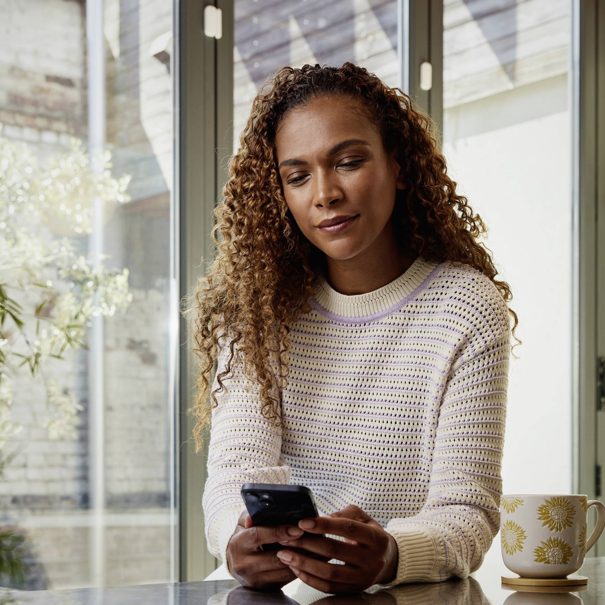 Eine Person mit lockigem Haar sitzt an einem Tisch und hält ein Smartphone in der Hand, blickt nachdenklich darauf. Im Hintergrund ist ein Fenster zu sehen.