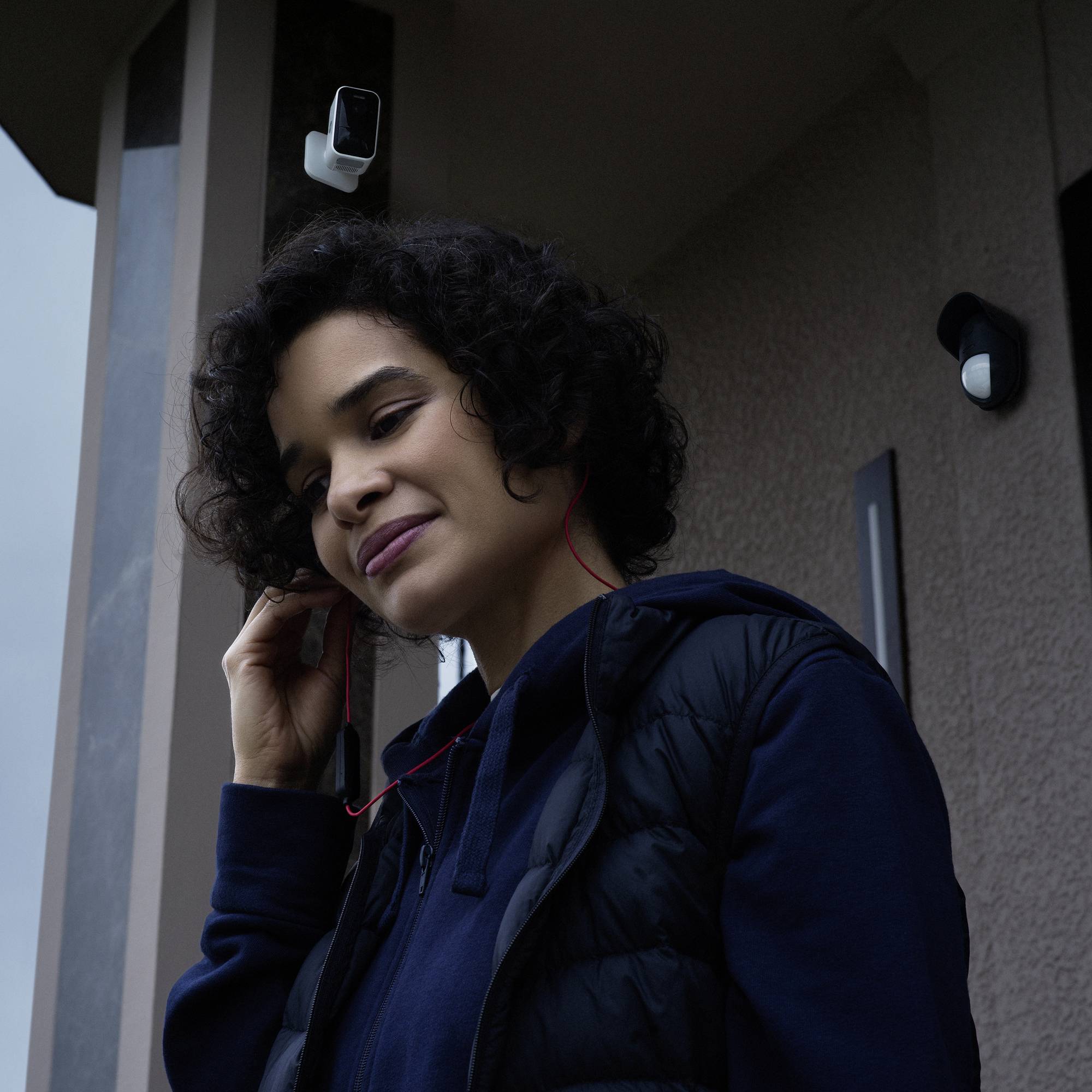 Frau mit Kopfhörern, lächelnd nach unten schauend, steht unter einem Dachvorsprung, der mit einer Überwachungskamera ausgestattet ist.