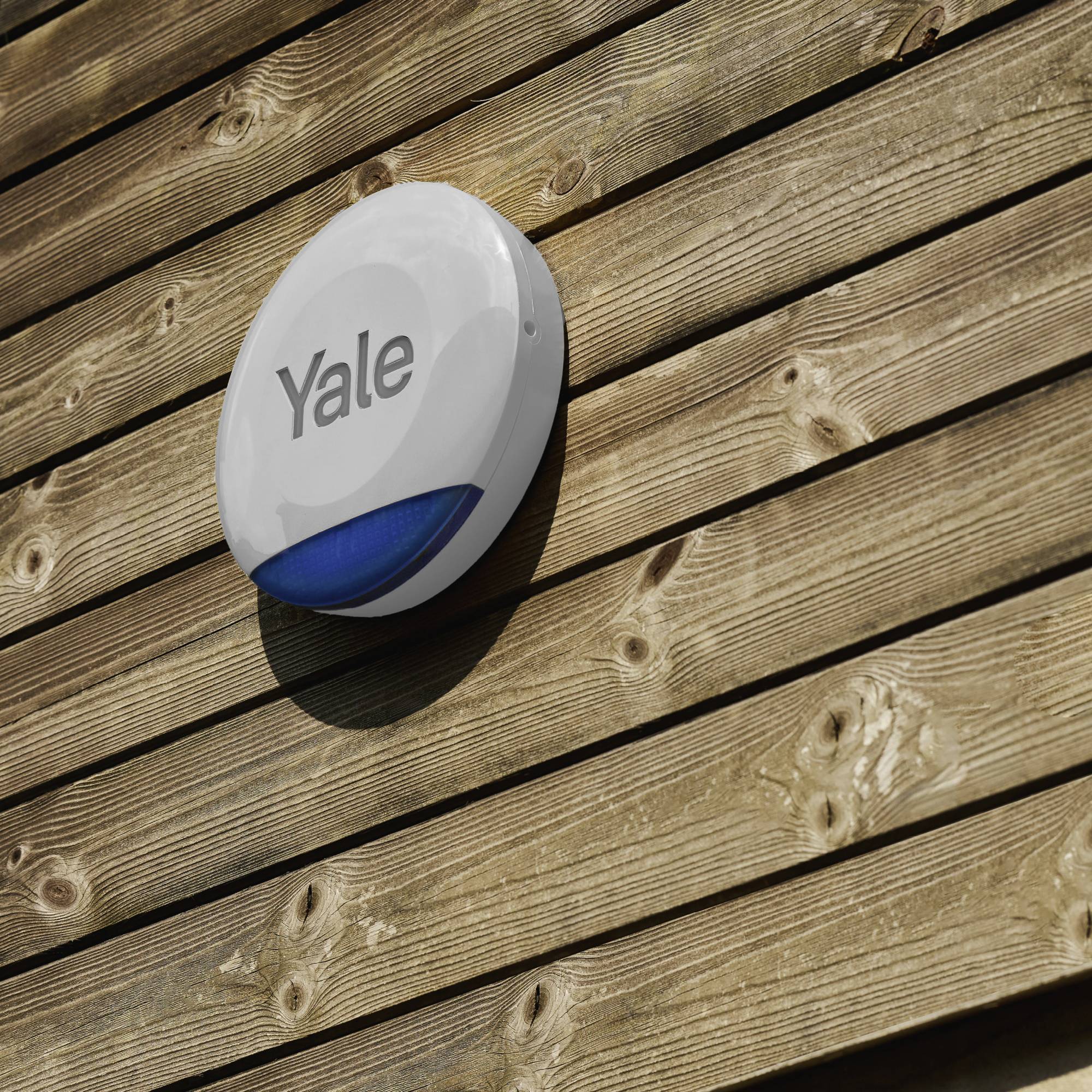 Hauswand mit einer kreisförmigen Alarmanlage der Marke Yale. Sie ist an einer Holzverkleidung angebracht.