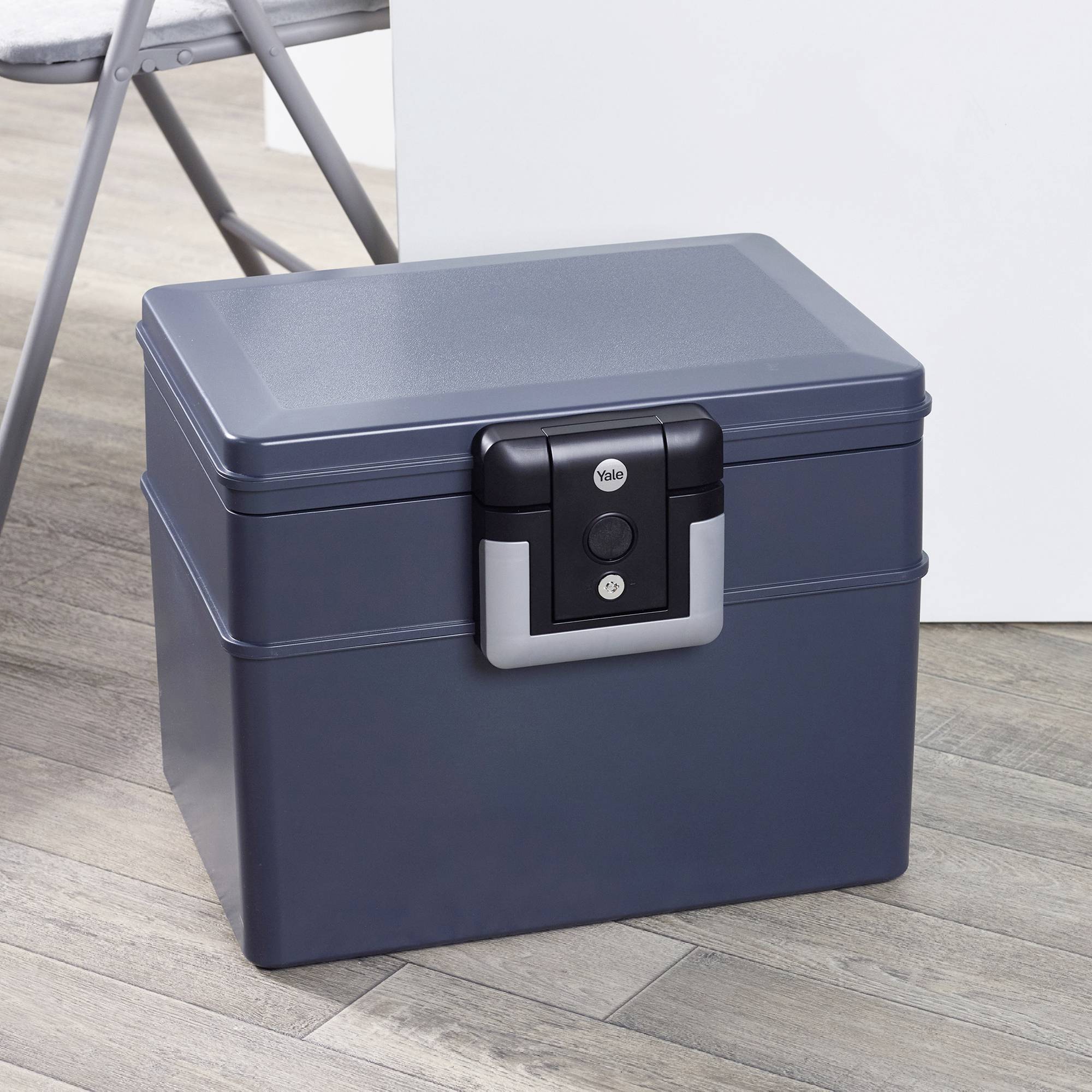 Eine schwarze, verschließbare Box steht auf einem Holzboden neben einem weißen Tisch und einem Klappstuhl.