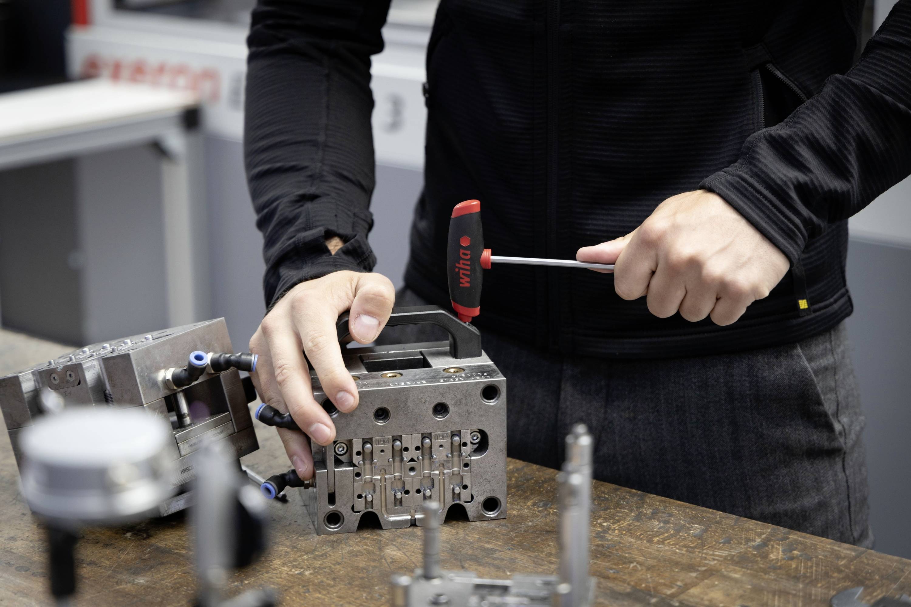Eine Person bearbeitet eine Metallform mit einem Hammer auf einem Tisch, auf dem weitere Werkzeuge liegen. Sie trägt einen schwarzen Pullover.