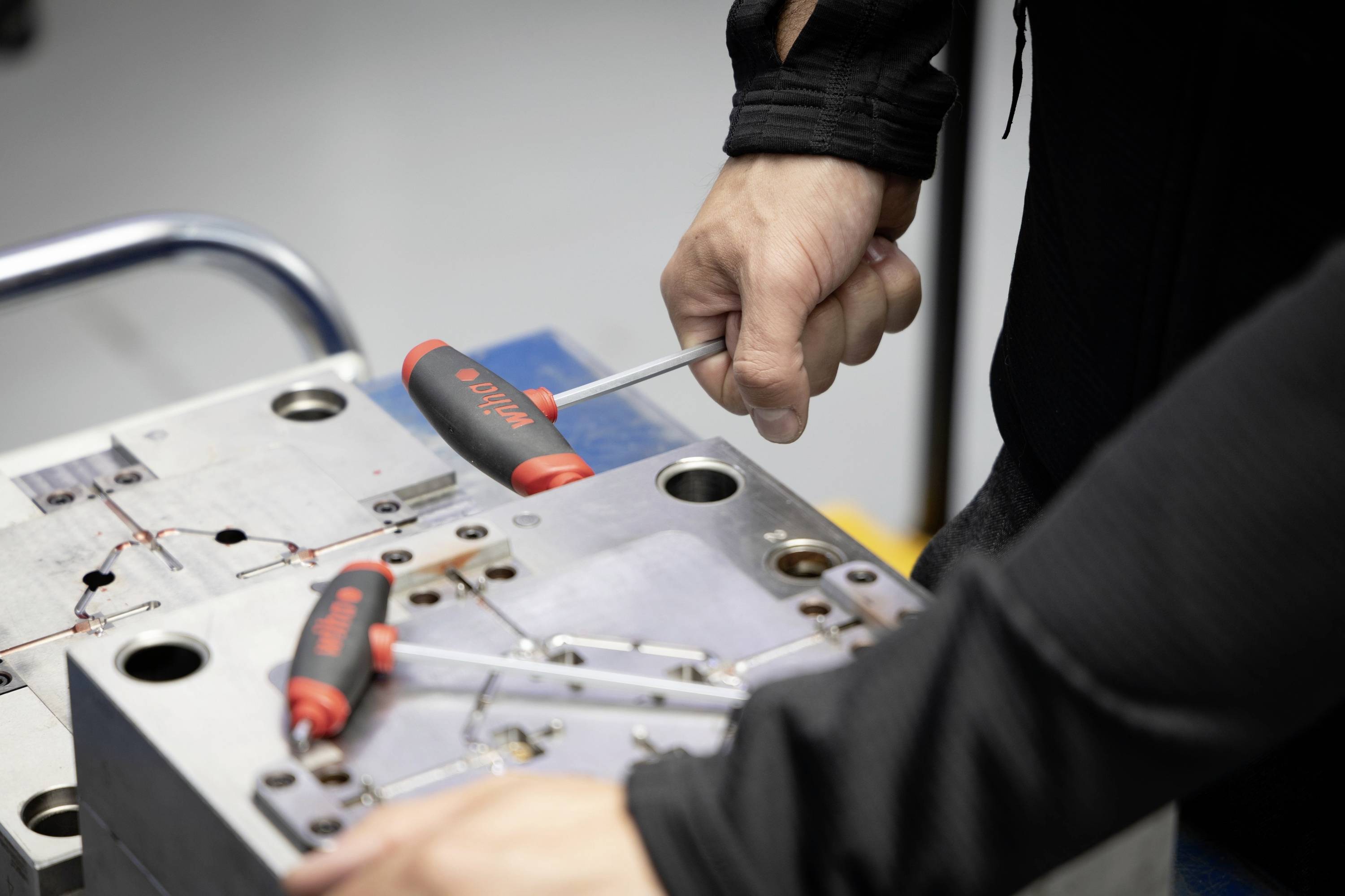 Eine Person zieht mit Schraubendrehern an einer Metallplatte in einer Werkstatt. Zwei rote Schraubendreher sind sichtbar.