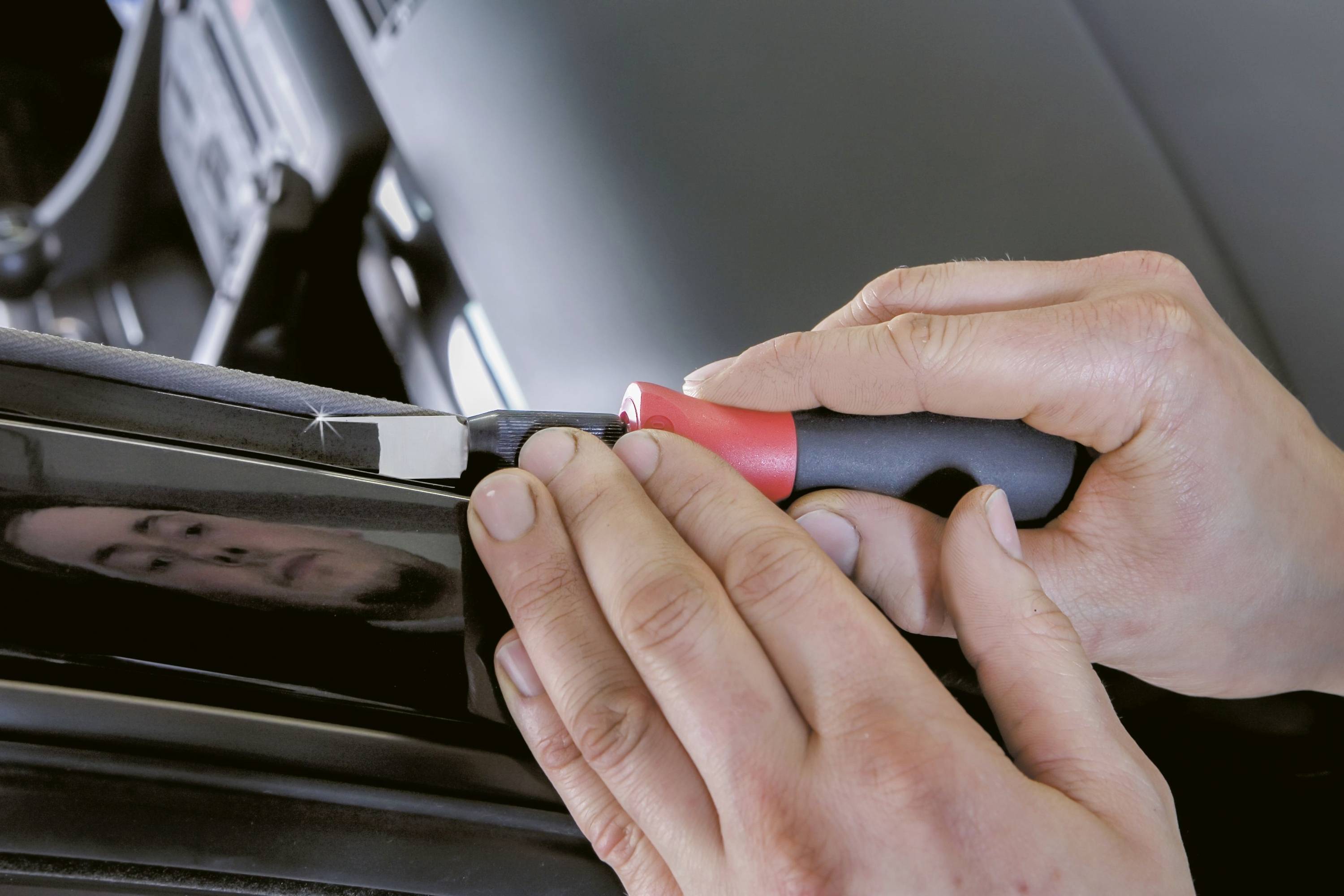 Zwei Hände verwenden ein Werkzeug, um am Rand eines Fahrzeugs zu arbeiten. Die Szene zeigt präzise, manuelle Arbeit an einem Auto.