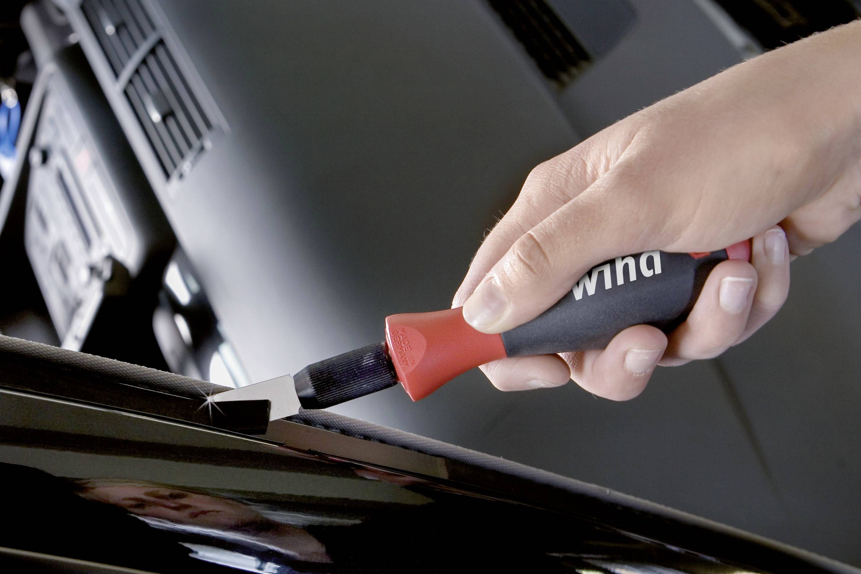 Eine Hand hält einen rot-schwarzen Schraubenzieher an der Kante eines schwarzen Objekts. Im Hintergrund sind unscharfe Geräte zu sehen.