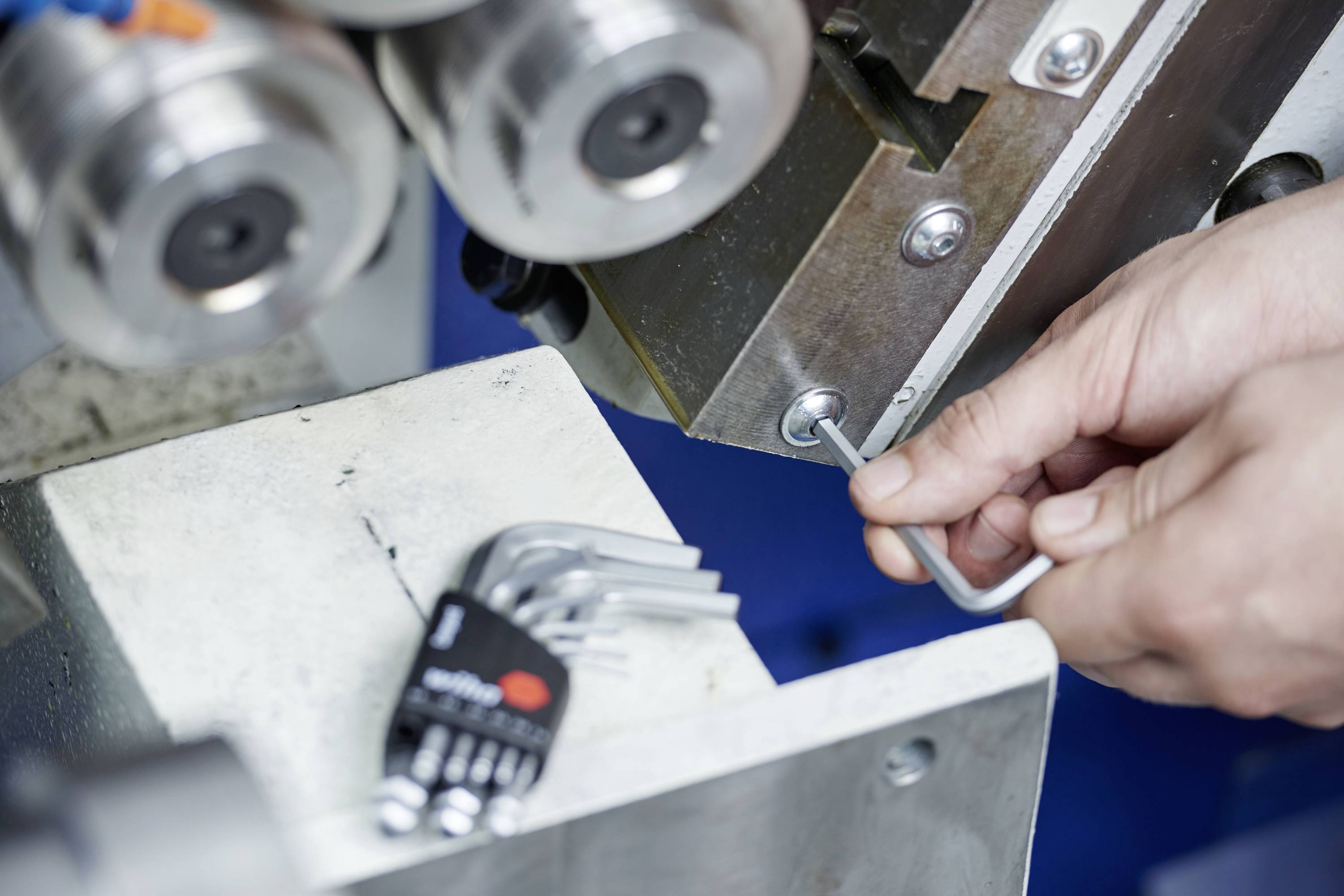 Eine Person zieht eine Schraube an einer Maschine mit einem Inbusschlüssel an. Daneben liegt ein Satz Inbusschlüssel.