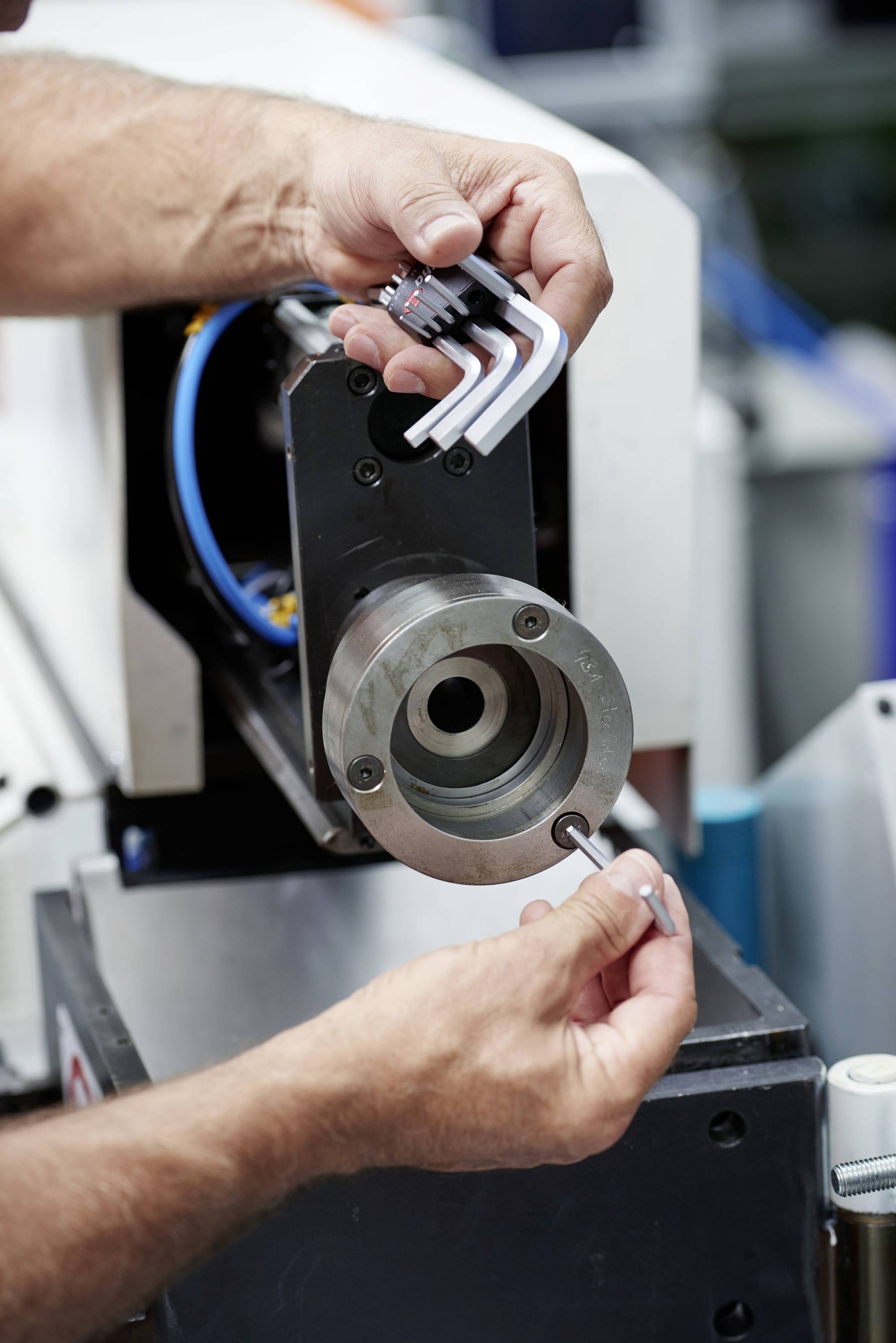 Eine Person justiert eine Metallmaschine mit einem Inbusschlüssel, um Teile festzuziehen. Im Hintergrund sind Teile der Werkstatt sichtbar.