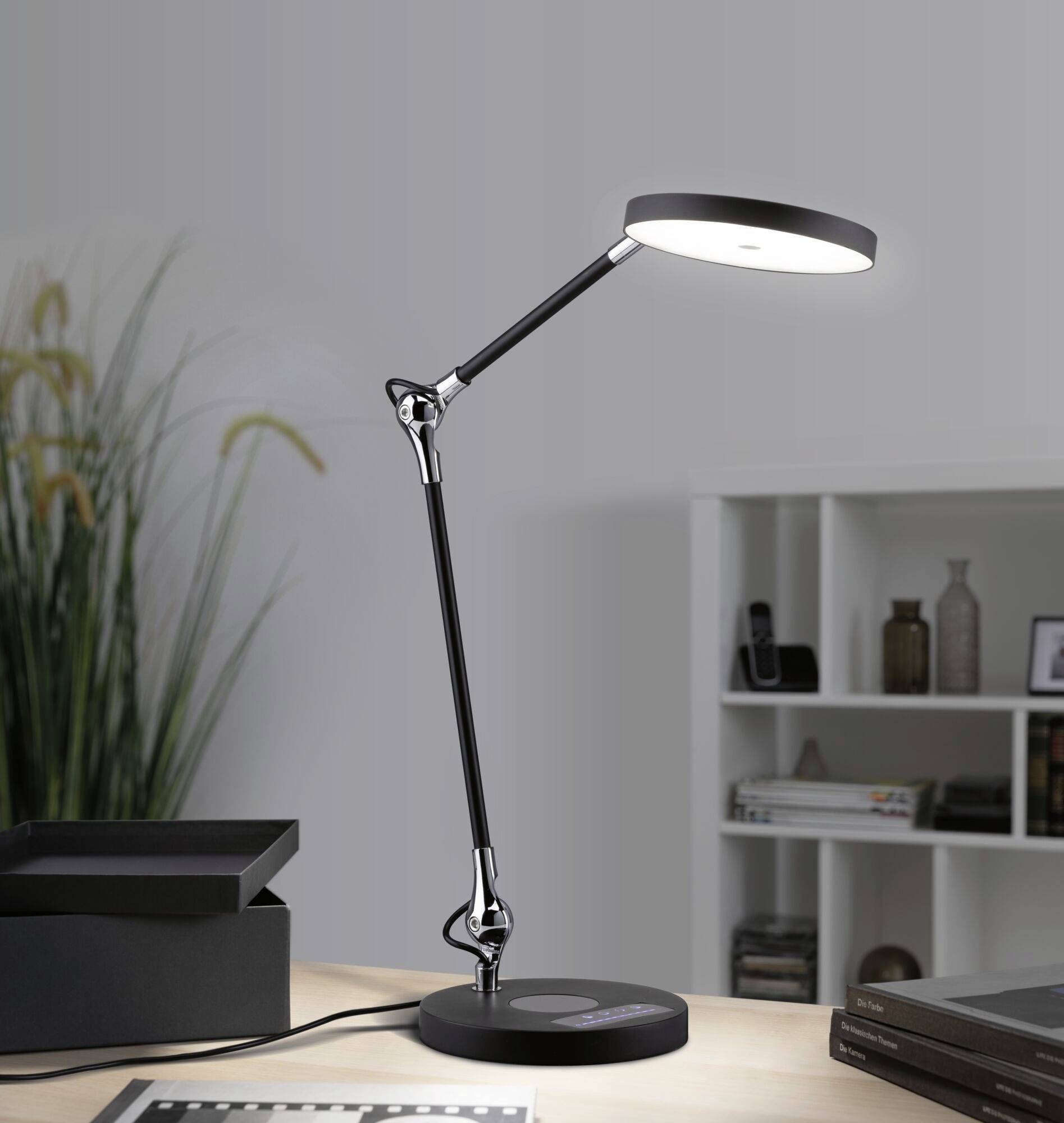 Eine moderne, schwarze Schreibtischlampe mit verstellbarem Arm, beleuchtet einen Tisch. Im Hintergrund sind ein Regal und Pflanzen sichtbar.