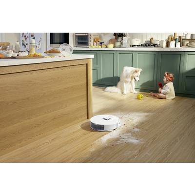 Ein Haushaltsroboter reinigt Mehl auf einem Holzboden. Im Hintergrund spielt ein Kleinkind mit einem Hund in der Küche.