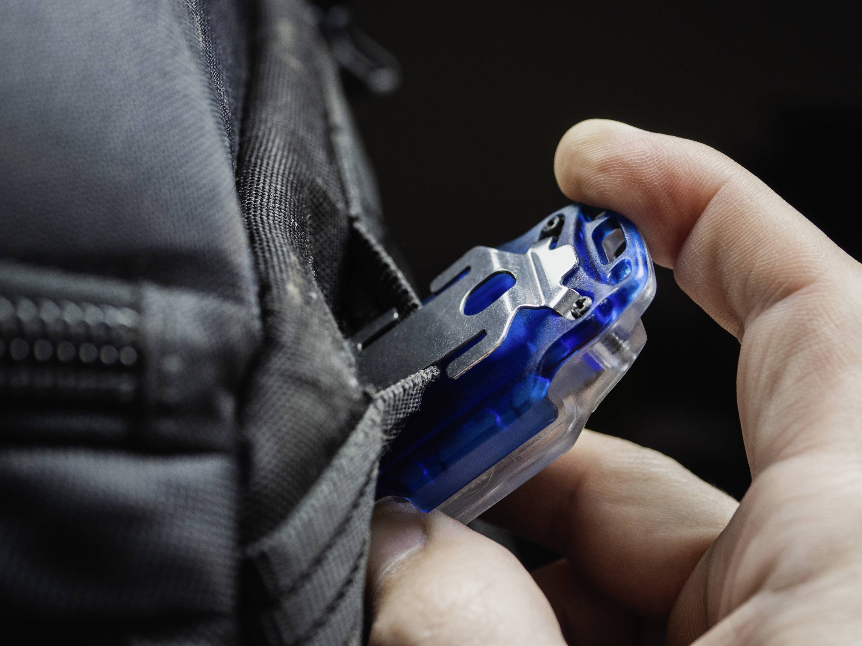 'Hand befestigt einen blauen Communicator an einem schwarzen Rucksack. Kommuniziert vermutlich über Funk oder digital, Zusammenhang unklar.'