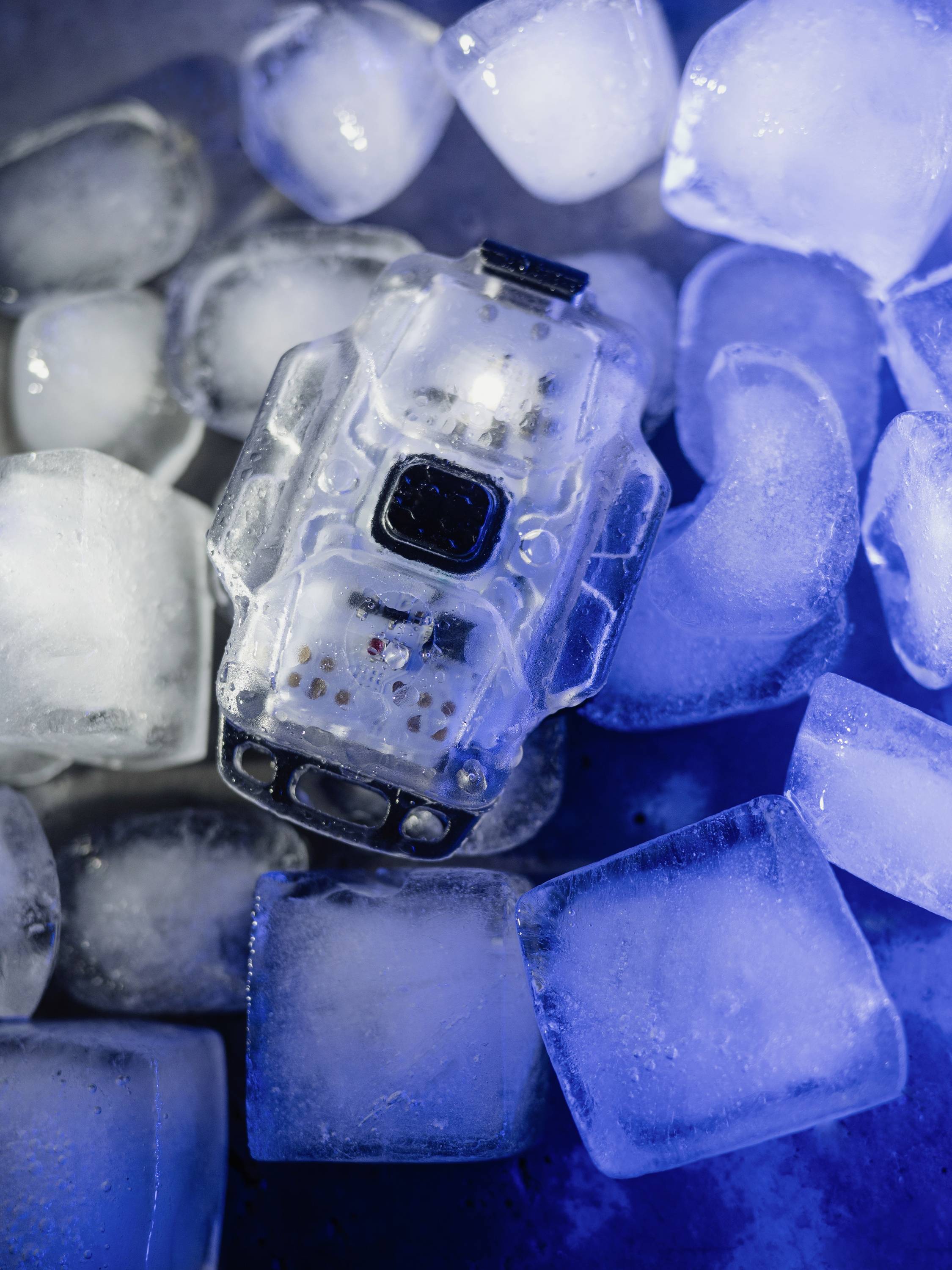 Ein elektronisches Gerät in transparentem Gehäuse liegt zwischen Eiswürfeln. Es ist von blauem Licht beleuchtet.