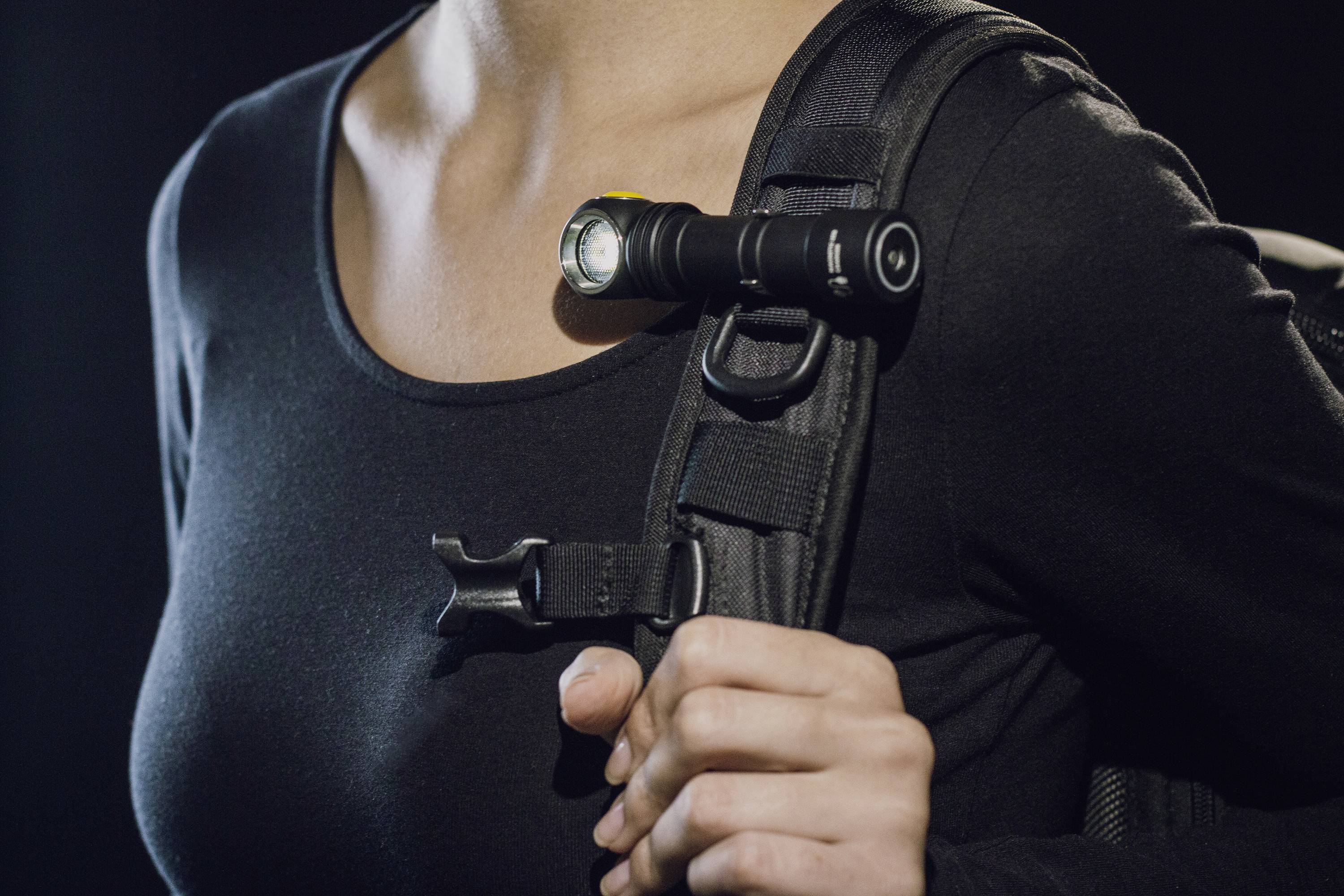 Person trägt einen Rucksack mit einer Taschenlampe, die an einem Schultergurt befestigt ist. Die Person hat ein dunkles Oberteil an.