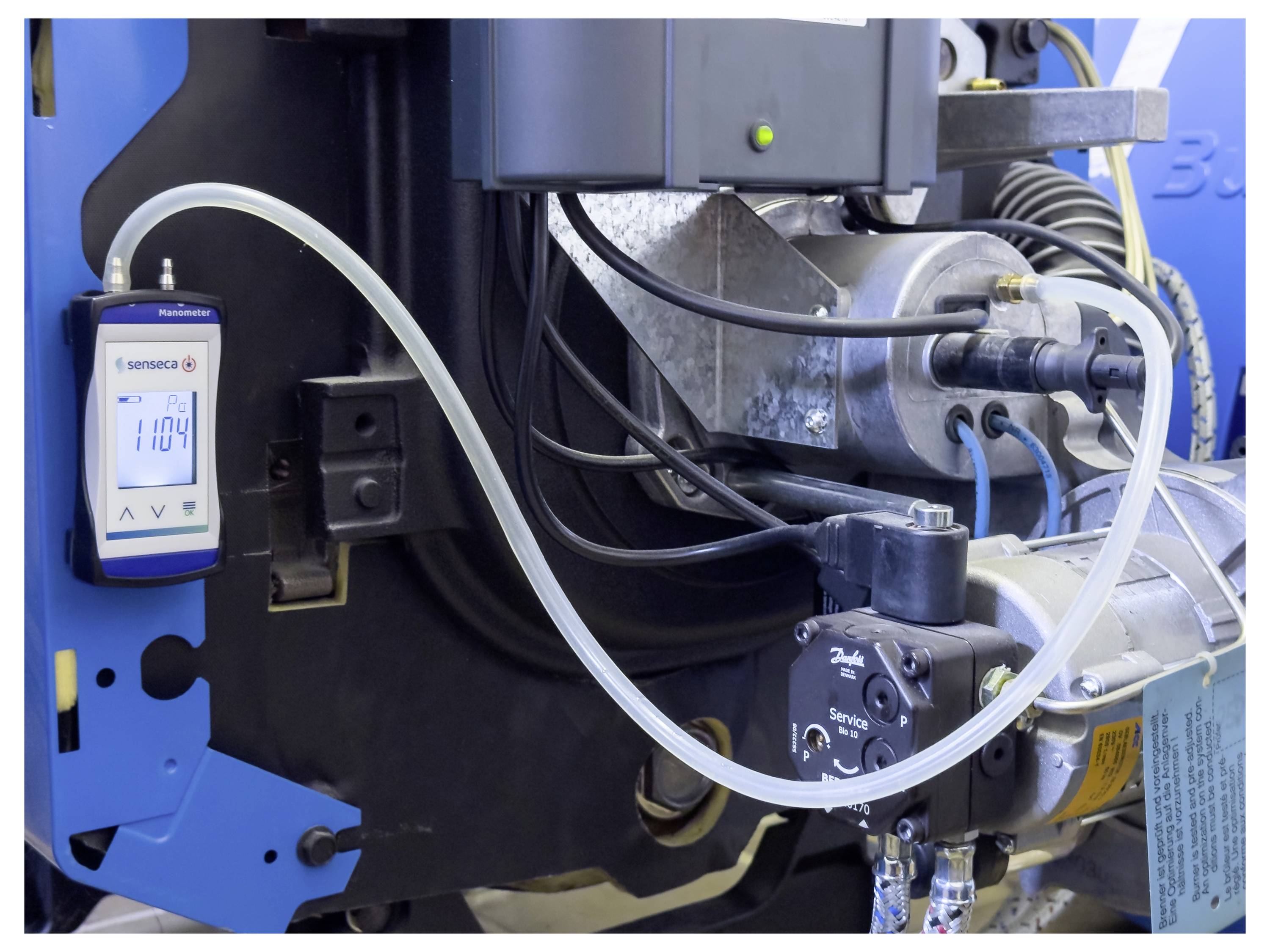 Eine technische Einrichtung, die ein Manometer mit der Anzeige '1104' zeigt, das mit Maschinen durch Kabel und Schläuche verbunden ist, möglicherweise Teil eines industriellen Systems.