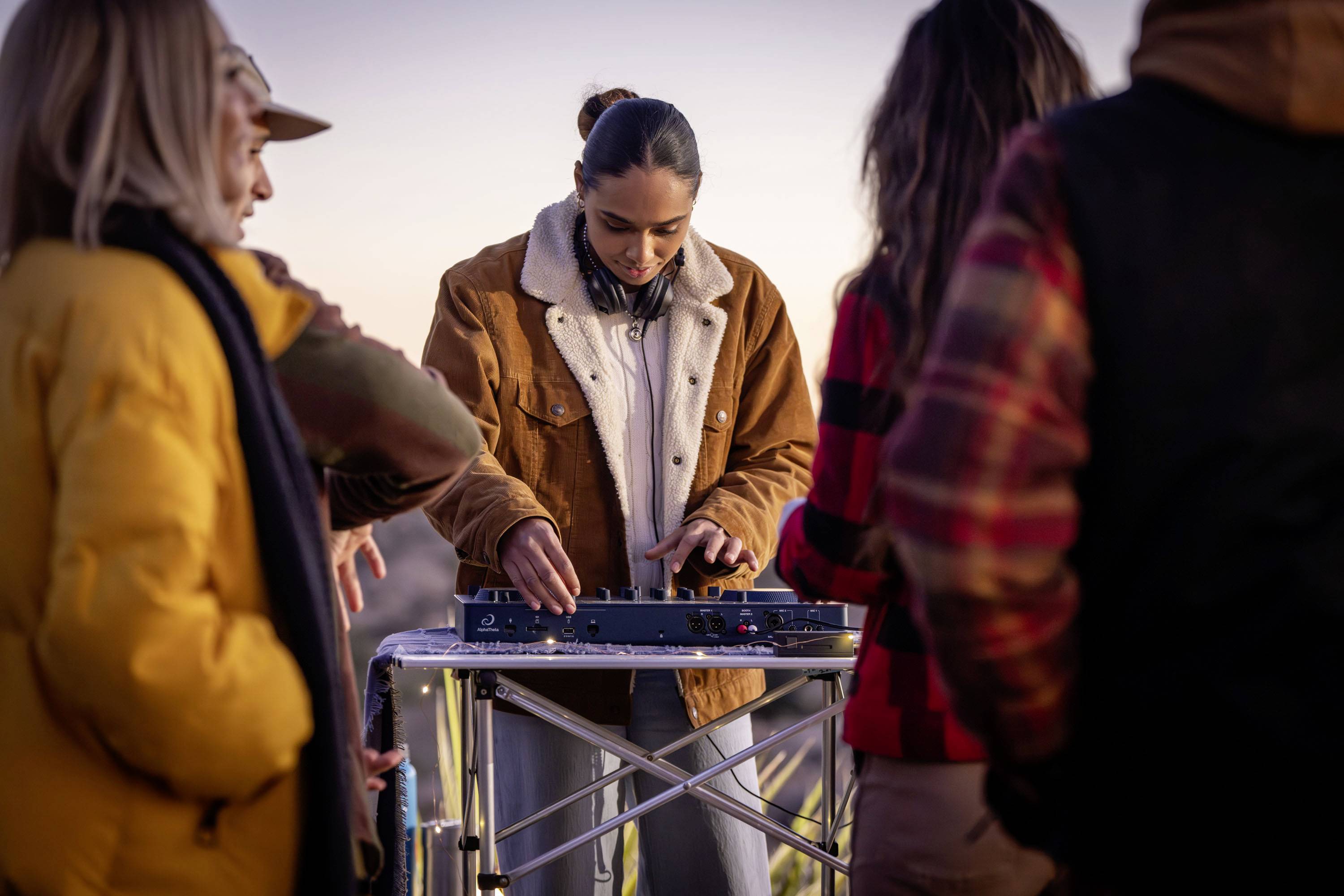 Eine Person mischt Musik an einem DJ-Controller im Freien, umgeben von anderen Menschen, die sich unterhalten. Es scheint Abend zu sein.