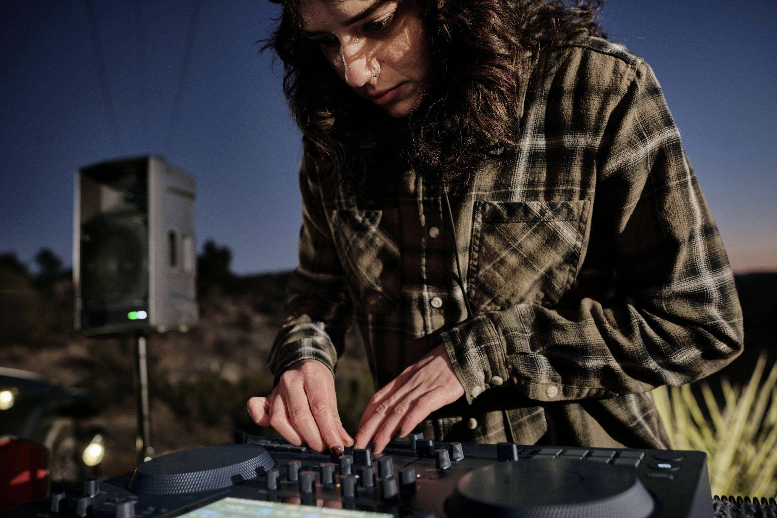 Frau mit lockigem Haar steht im Freien bei Dämmerung und mischt Musik auf einem DJ-Controller. Lautsprecher im Hintergrund.