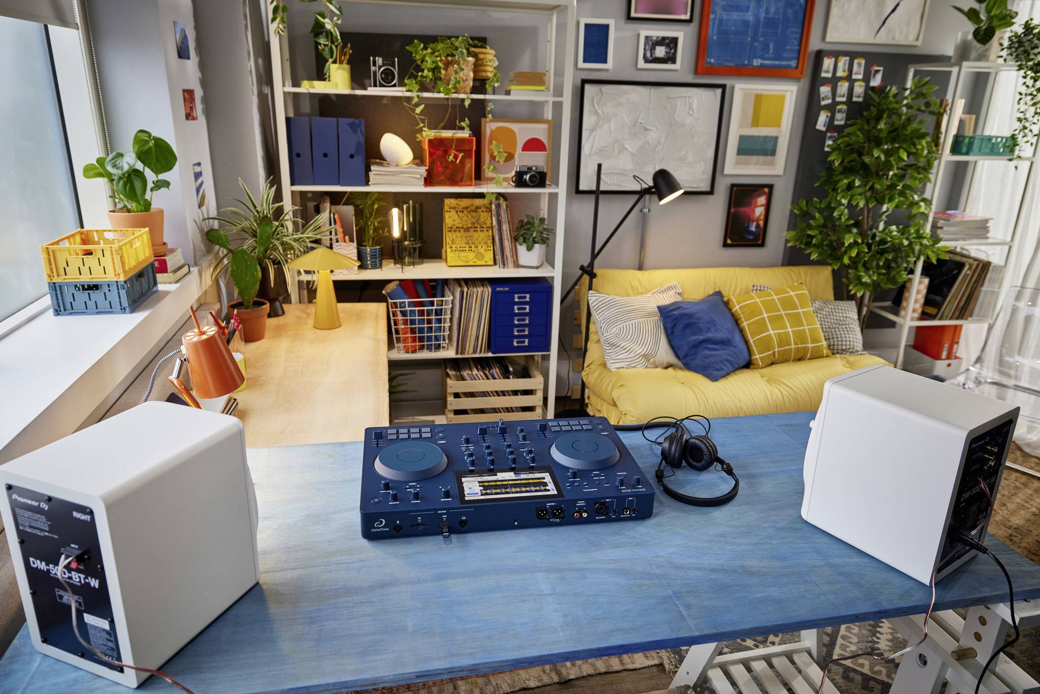 Ein Schreibtisch mit DJ-Mischpult und Kopfhörern. Im Hintergrund ein gemütliches Wohnzimmer mit gelbem Sofa und Bücherregalen.