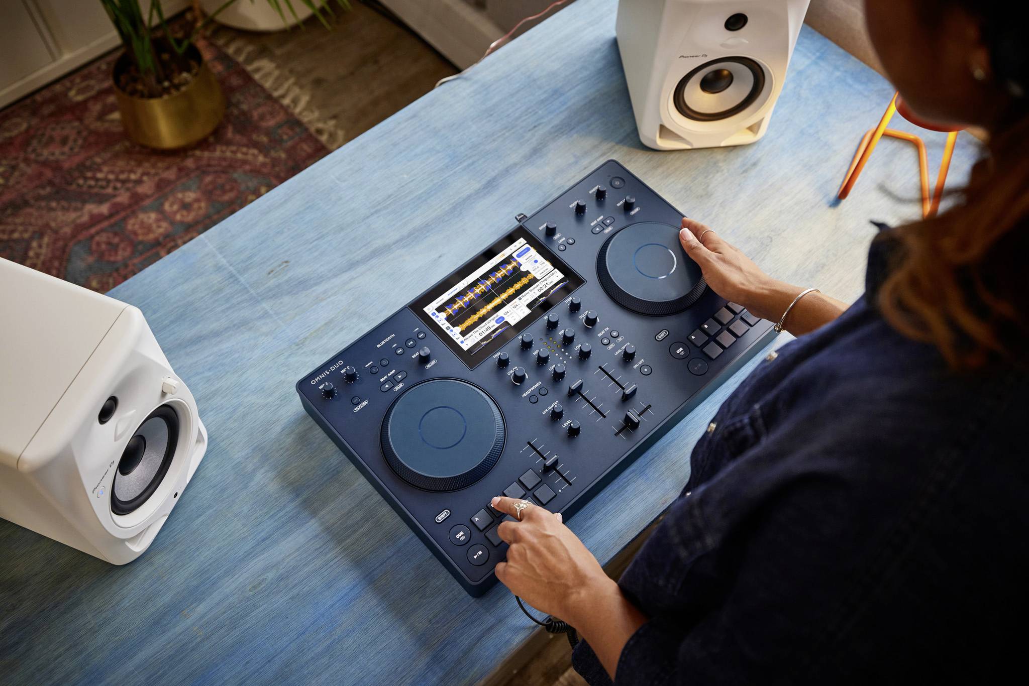 Eine Person bedient ein DJ-Mischpult mit einem Display, flankiert von zwei weißen Lautsprechern auf einem blauen Tisch.