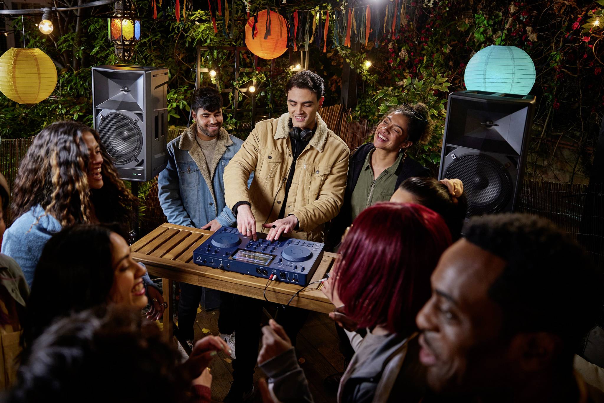 Eine Gruppe junger Menschen tanzt und lacht um einen DJ mit Kopfhörern, der draußen an einem Mischpult Musik auflegt. Bunte Laternen hängen herum.